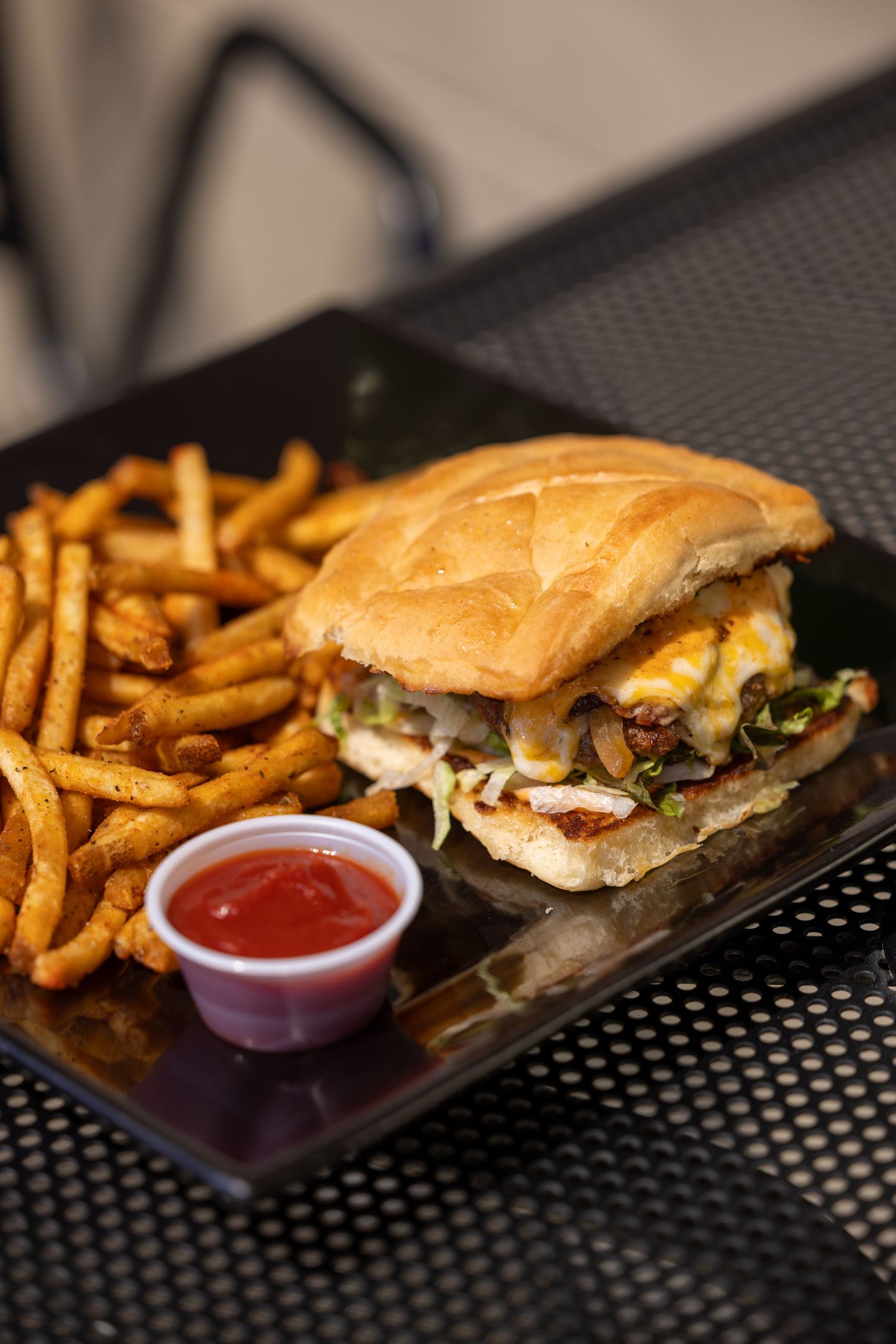 Juicy cheeseburger served on a black plate with crispy french fries and red dipping sauce.