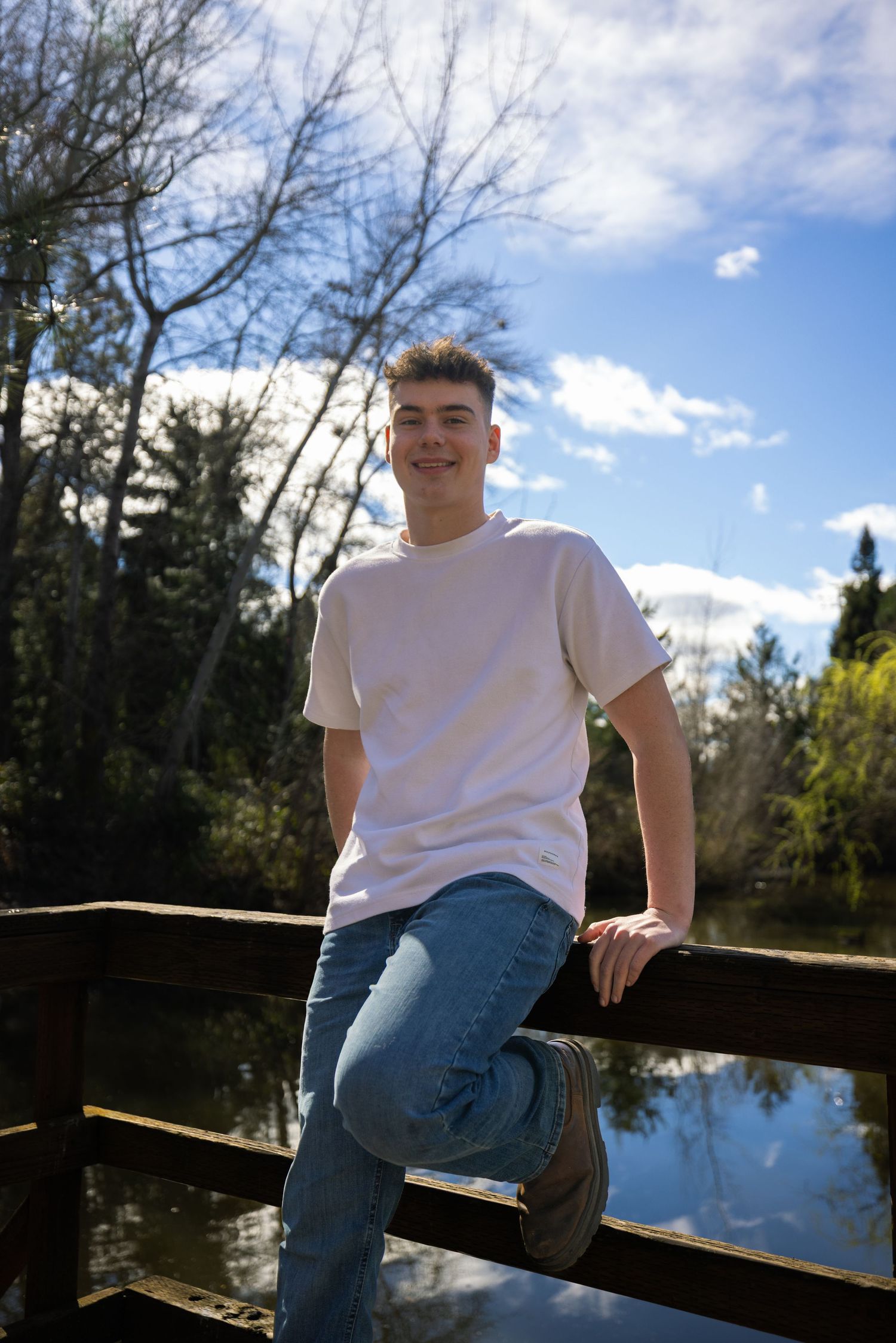 Young man in white t-shirt and jeans smiling on wooden bridge by pond surrounded by trees on sunny day.