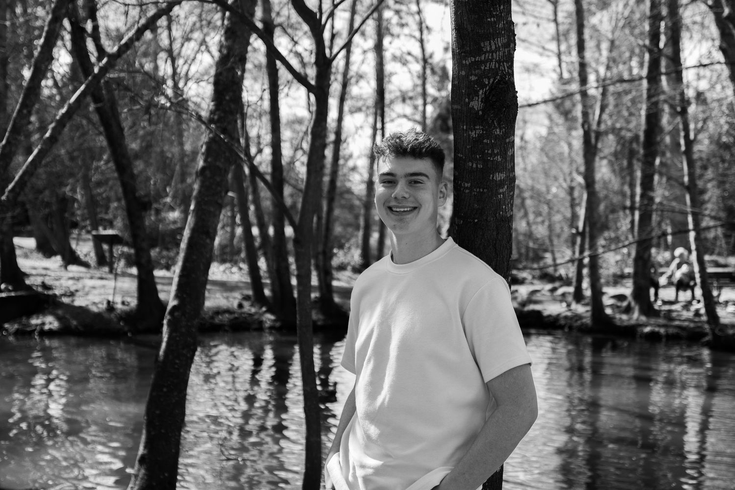 Young man smiling in white t-shirt standing by a forest stream in a black and white outdoor portrait photo.