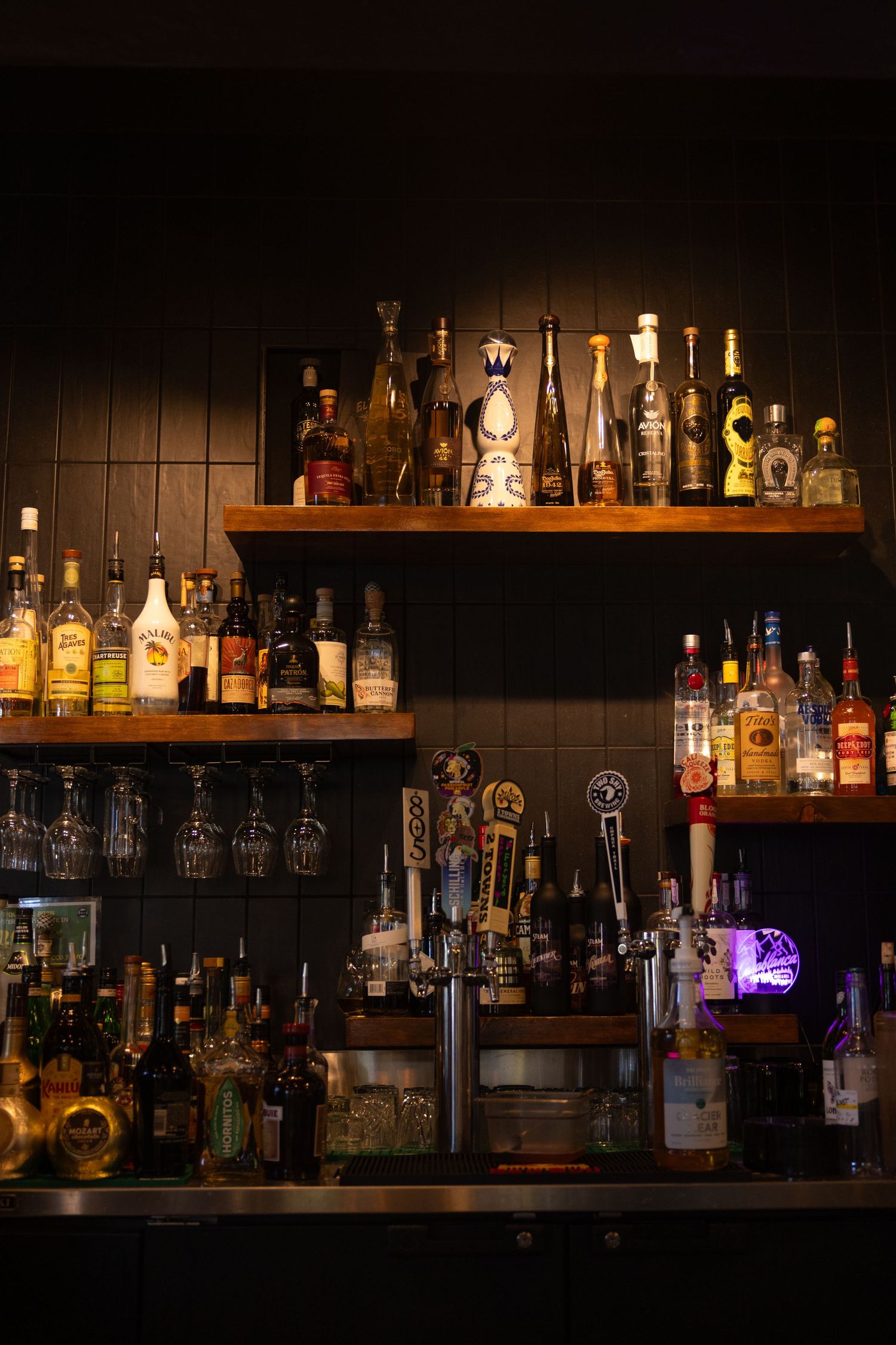 Illuminated bar shelves display various liquor bottles and glassware against a dark wall with warm ambient lighting.