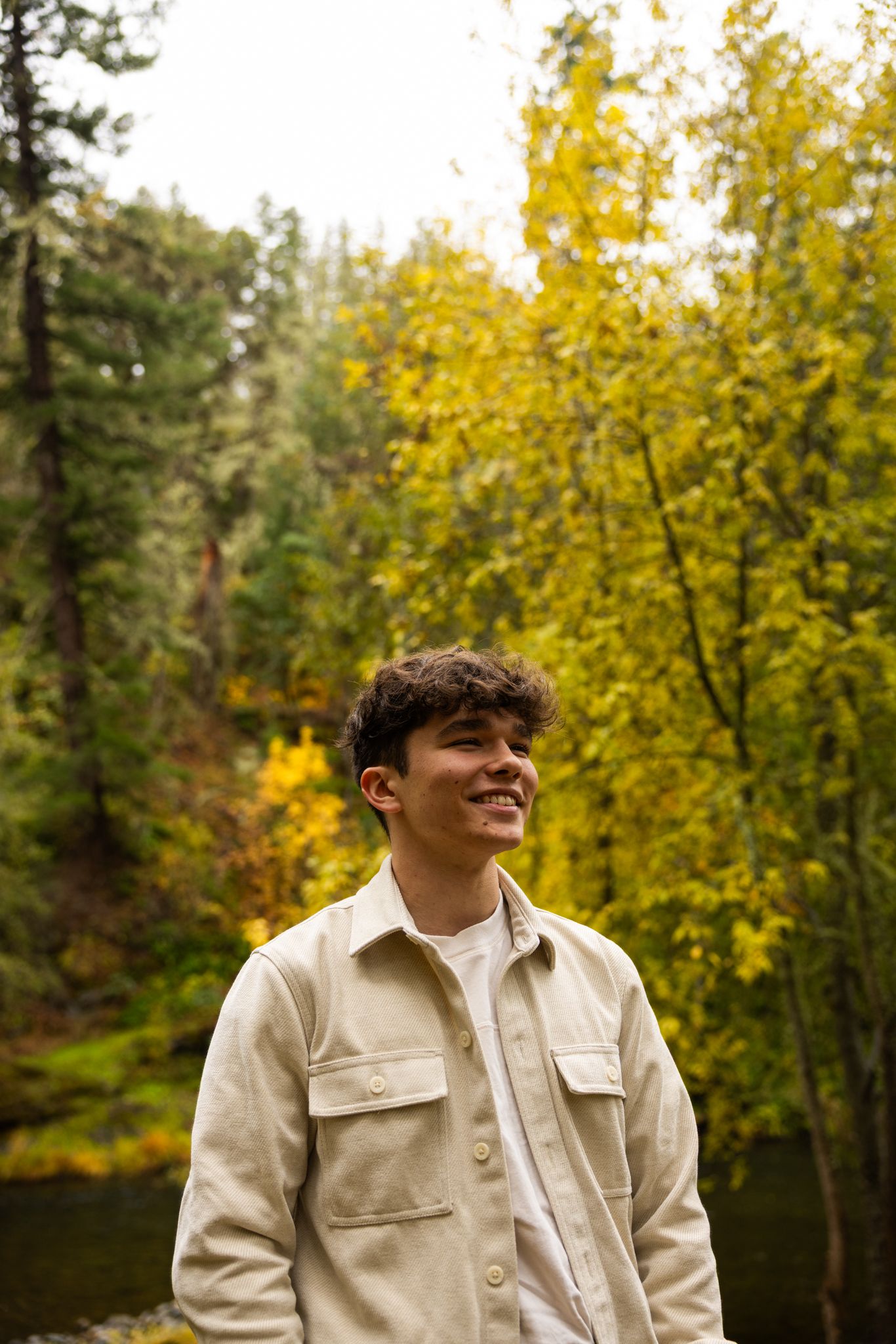 Smiling young man in beige jacket standing outdoors among vibrant yellow autumn trees in a forest setting.