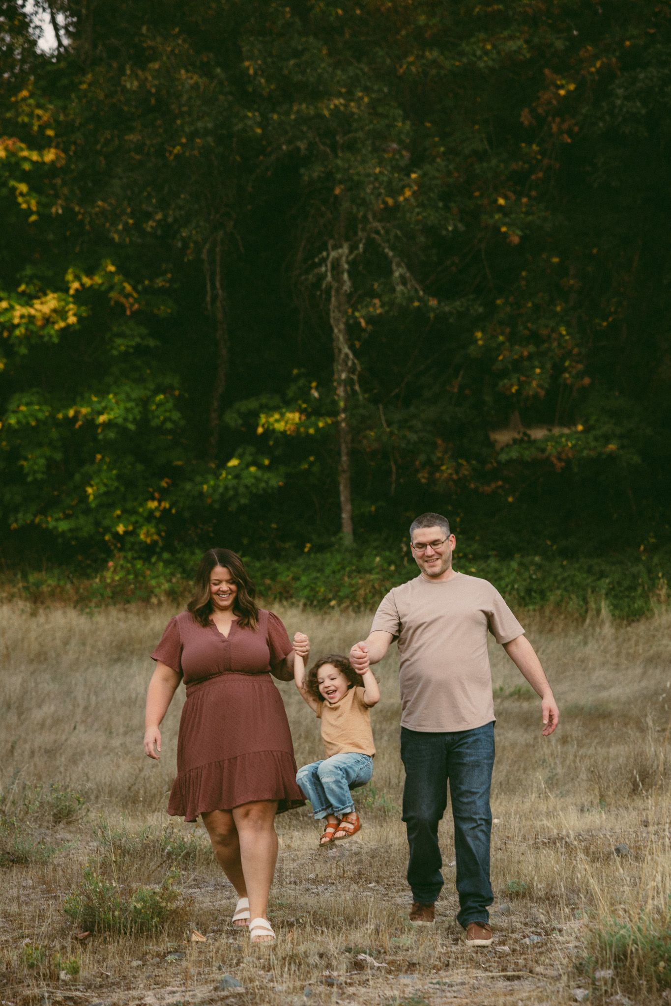 Family walks together through autumn field with trees and dried grass in background during golden hour.