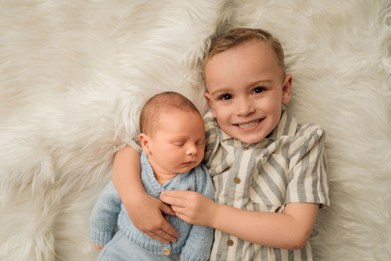Smiling toddler boy lying on a fluffy white rug with his sleeping newborn sibling.