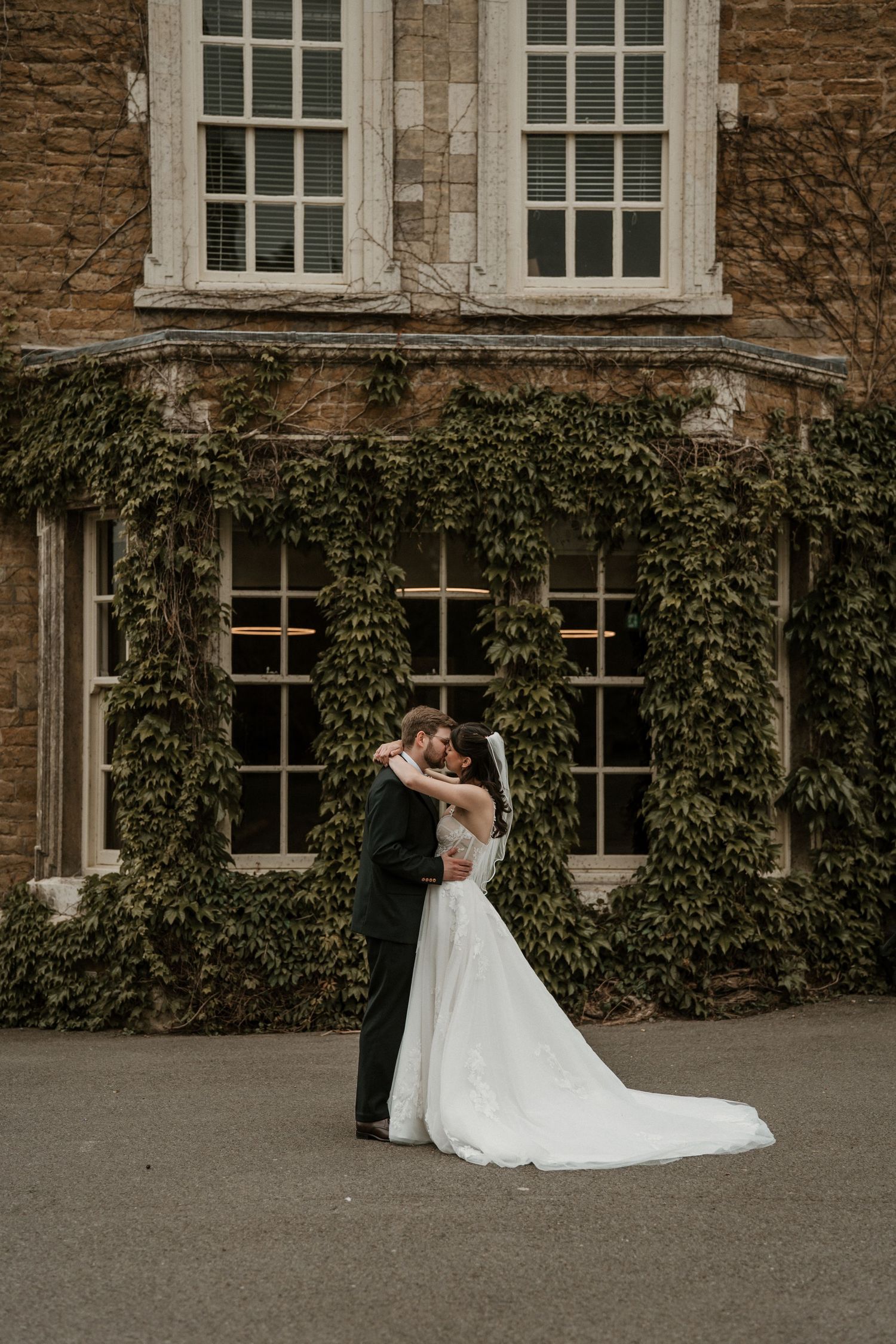 Bride and groom share a romantic embrace in front of an ivy-covered stone manor house on their wedding day.