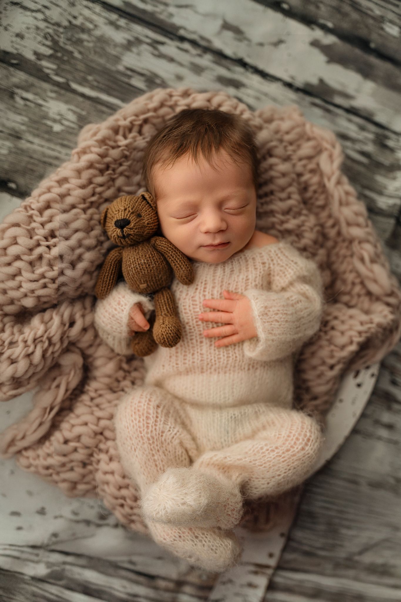 Sleeping newborn baby in cream knit outfit cuddling a brown teddy bear on a soft blanket.