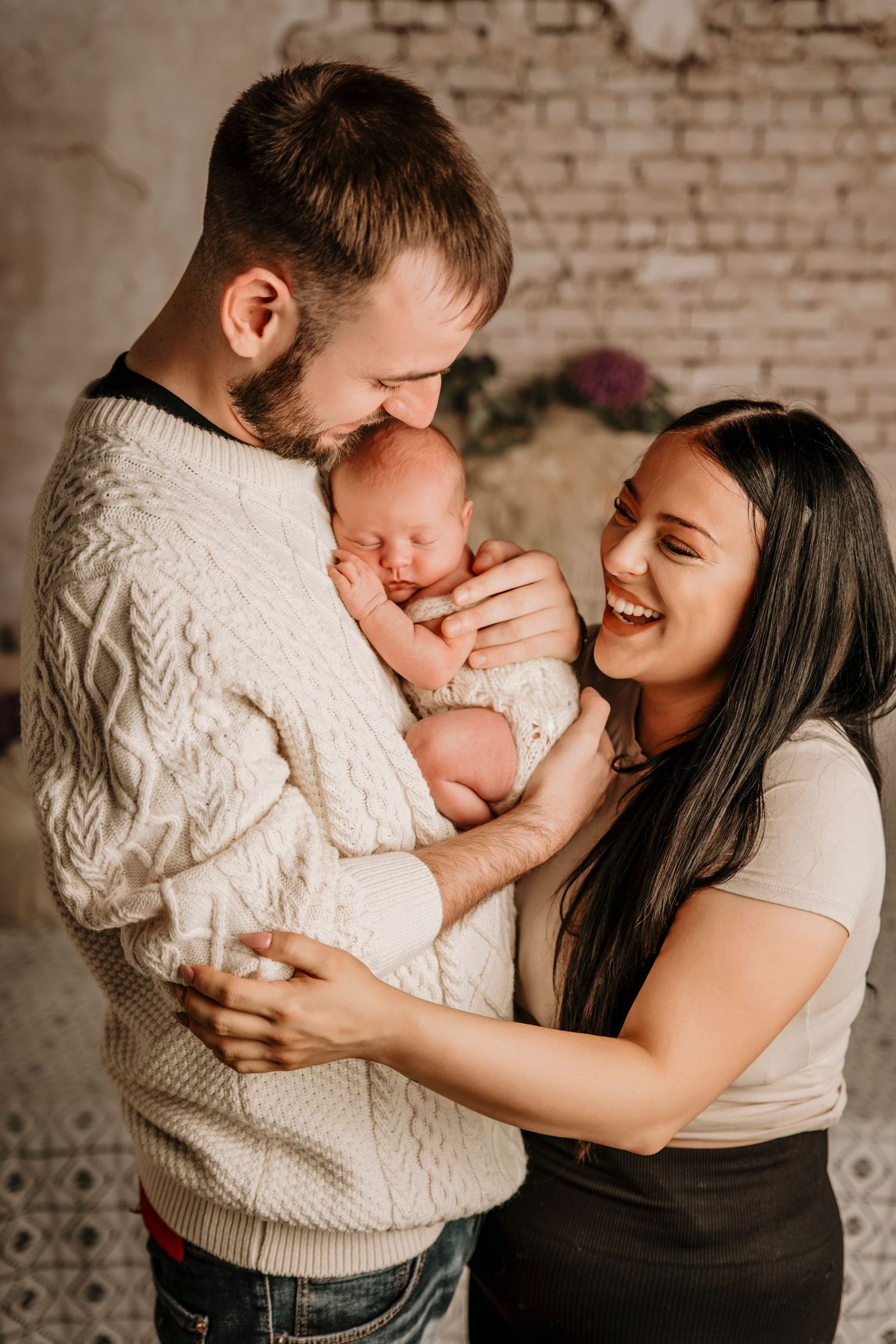 Happy young parents smiling and holding their sleeping newborn baby together in a cozy studio setting.