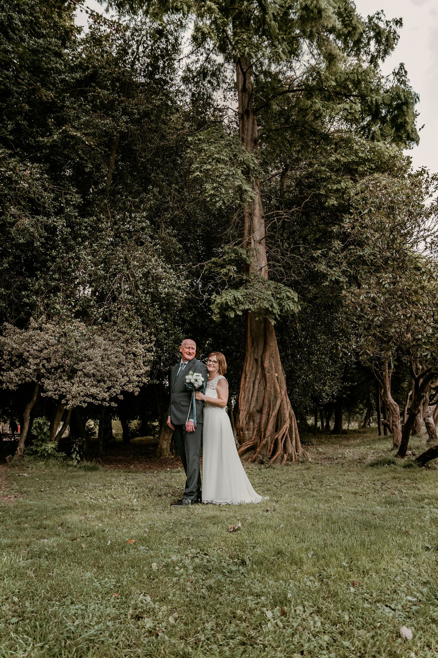 Bride and groom stand together in lush woodland garden with towering ancient trees on wedding day.