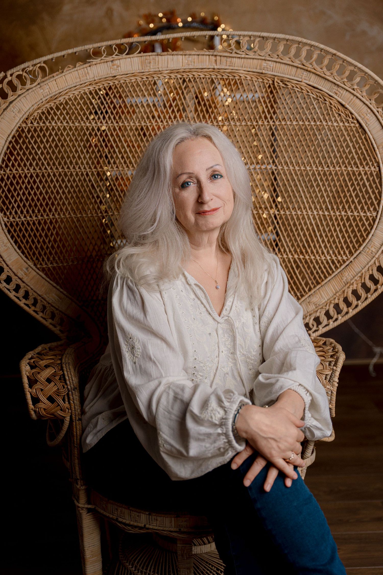 Elegant older woman with long gray hair sitting in a wicker peacock chair wearing a white blouse and jeans.