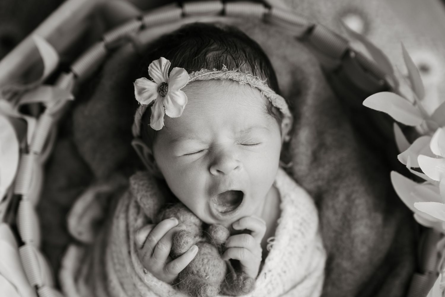 Black and white photo of yawning newborn baby girl with flower headband sleeping in a wicker basket.