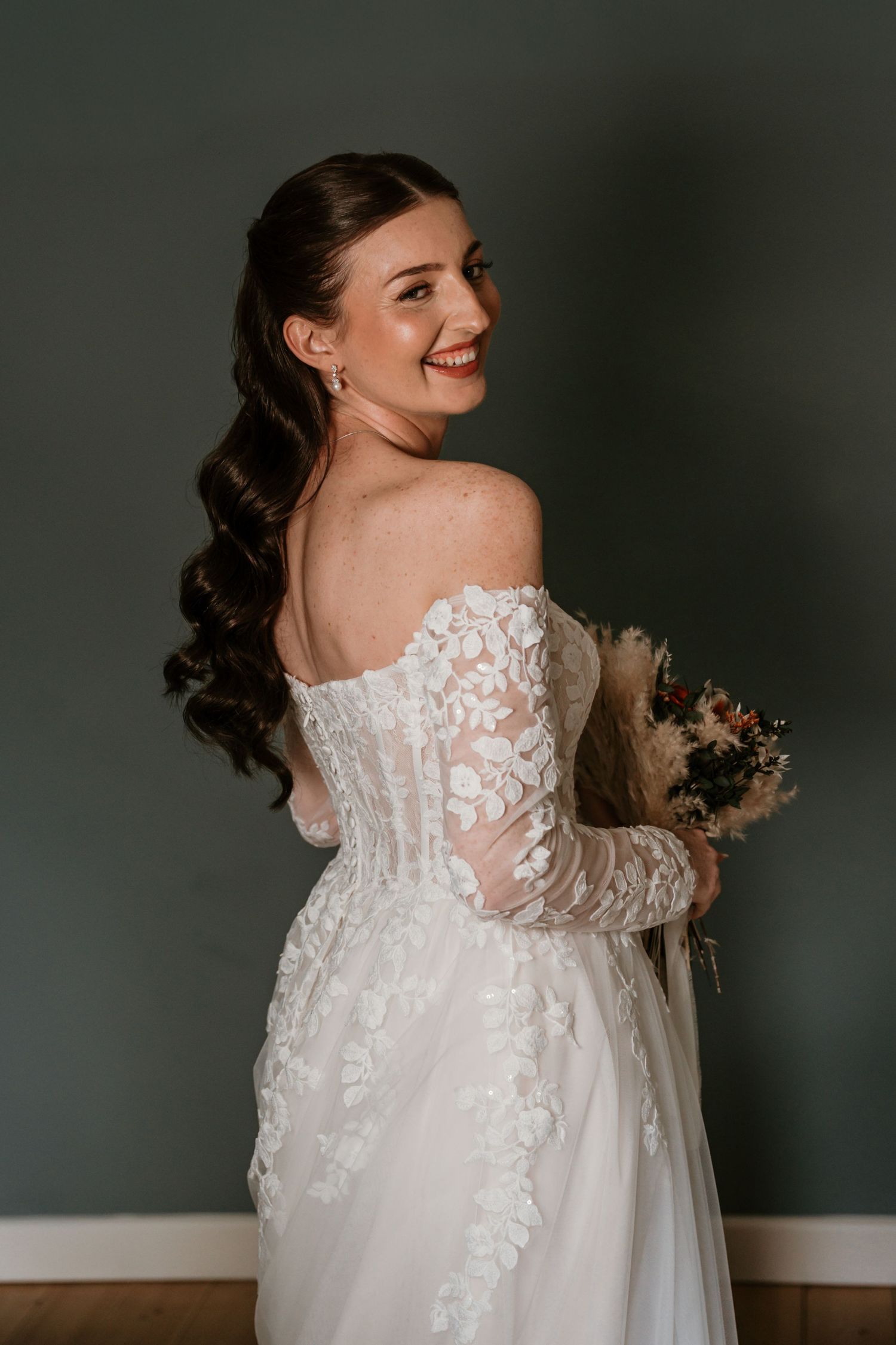 Smiling bride in an off-shoulder lace gown poses with her back to camera holding a dried flower bouquet.