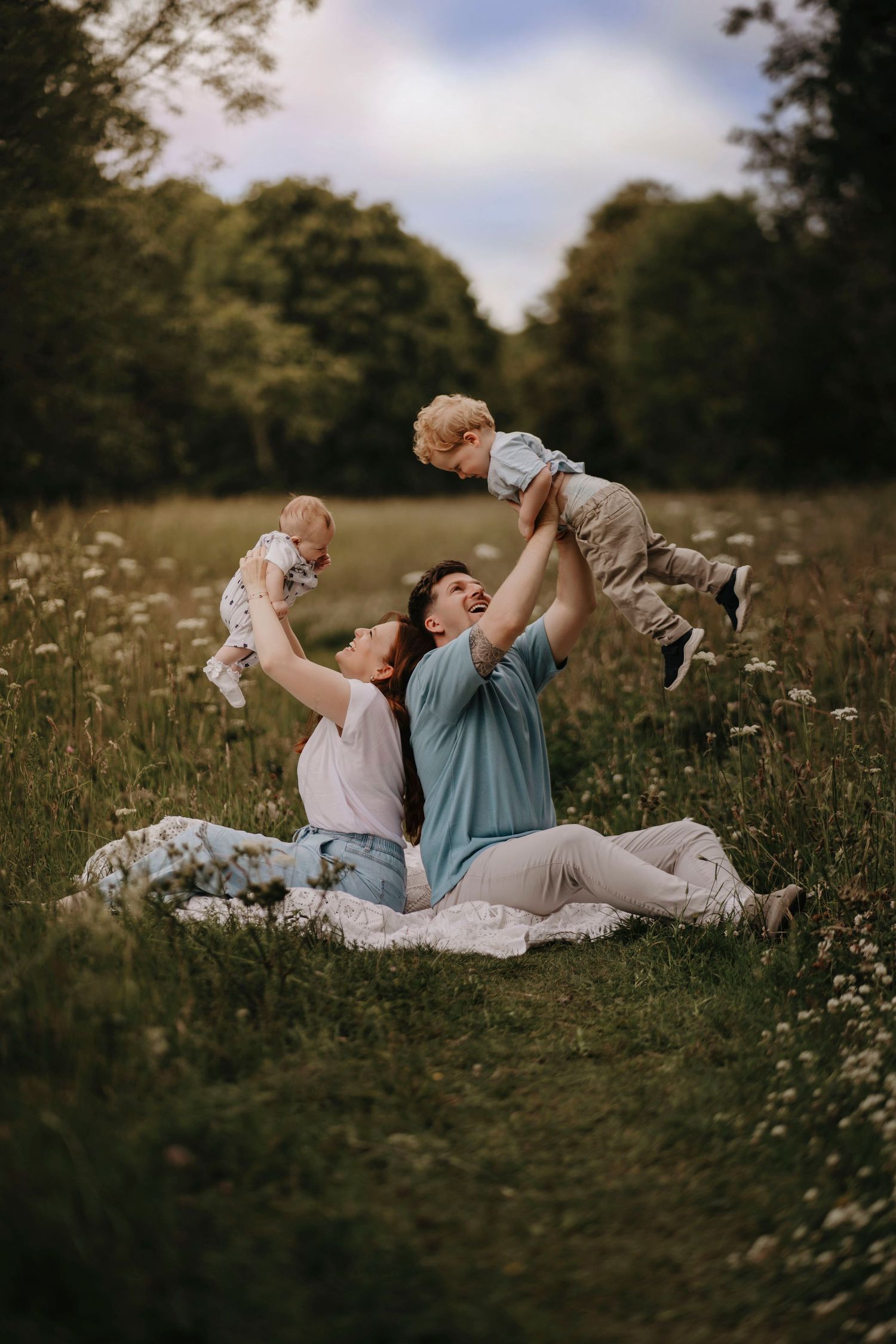 Family sitting in a wildflower meadow, joyfully lifting two young children into the air on a sunny day.