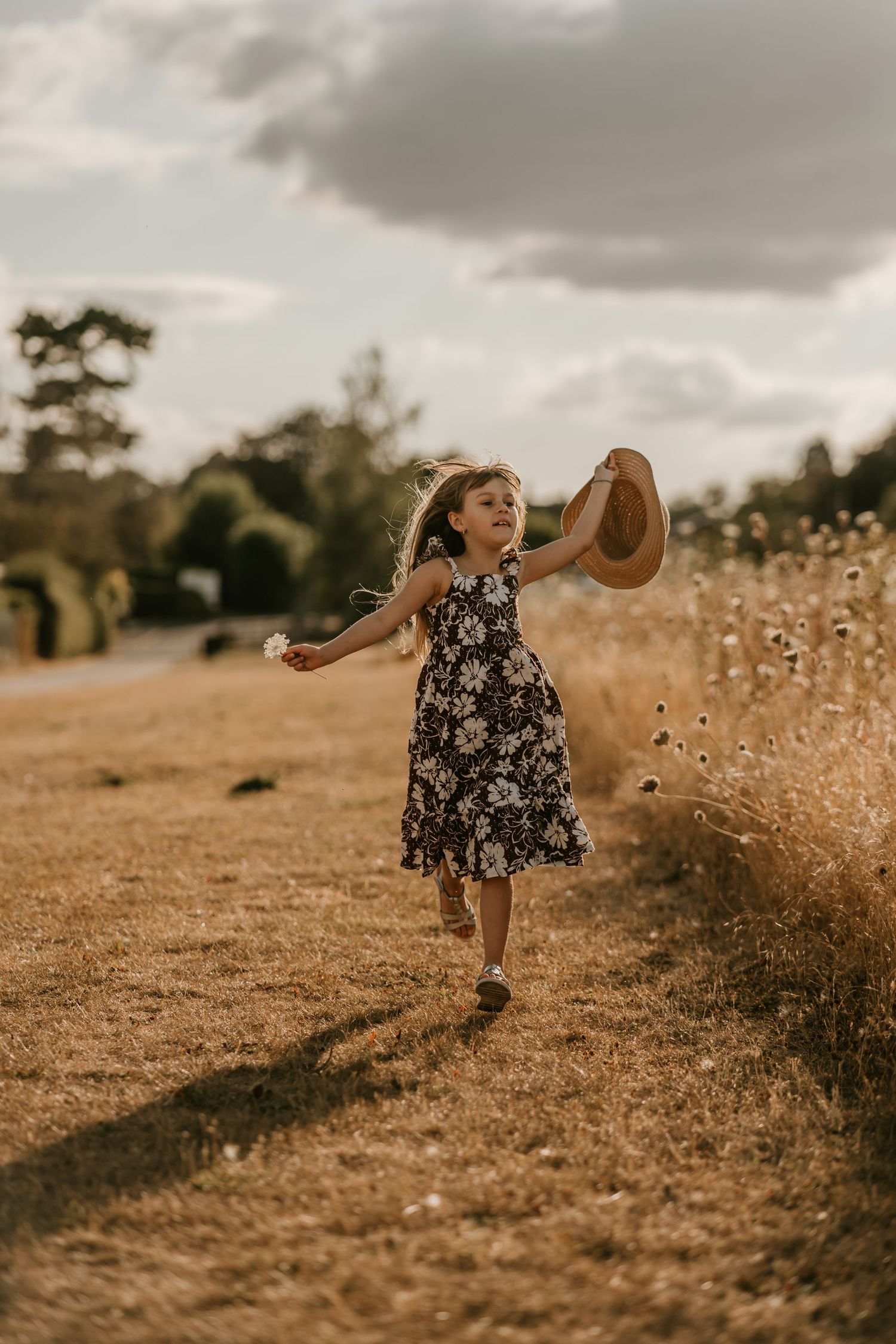 A joyful young girl in a floral dress runs through a golden field, holding a straw hat up in the air.