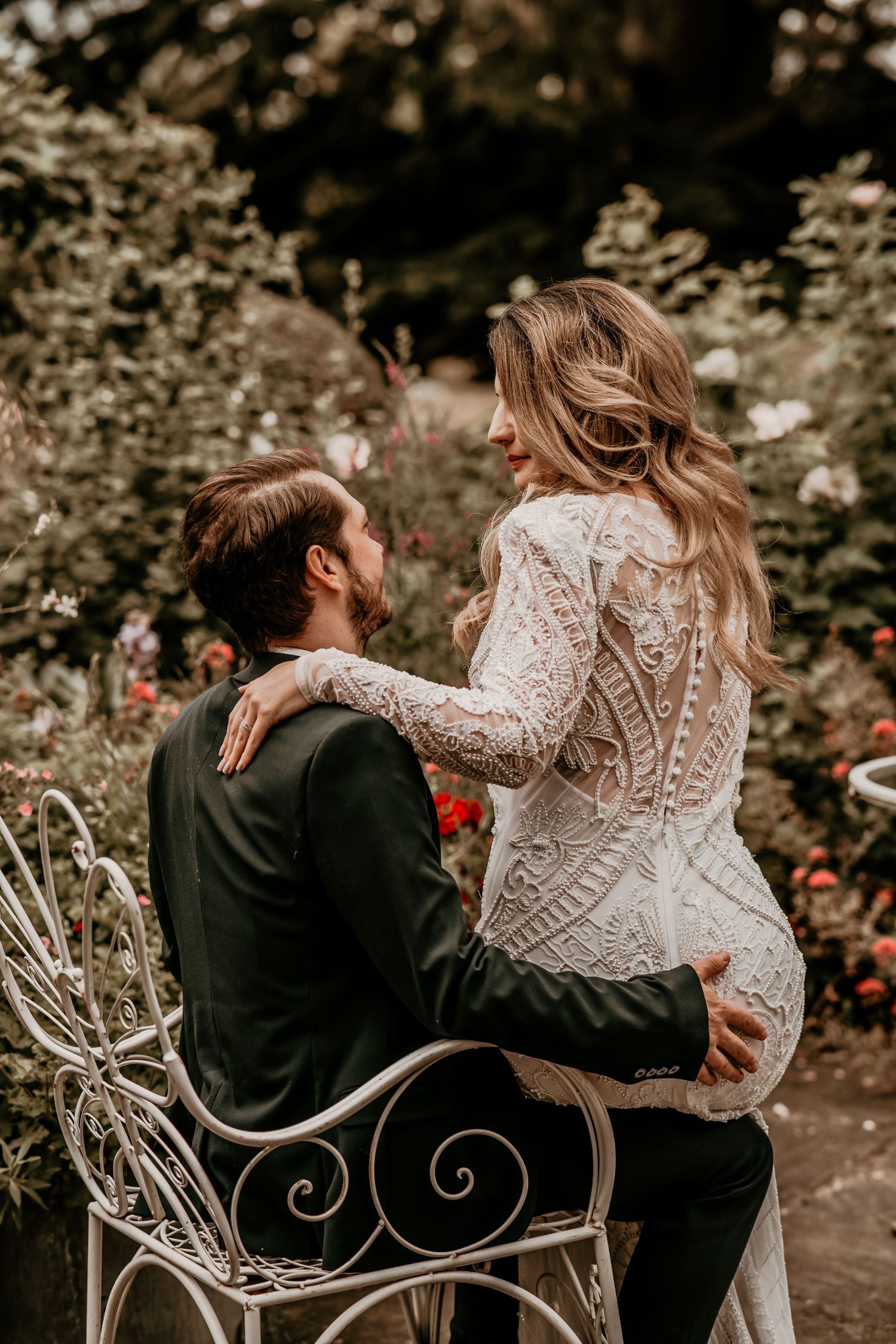Bride in long-sleeve lace gown sits with groom on ornate white bench surrounded by blooming rose garden.