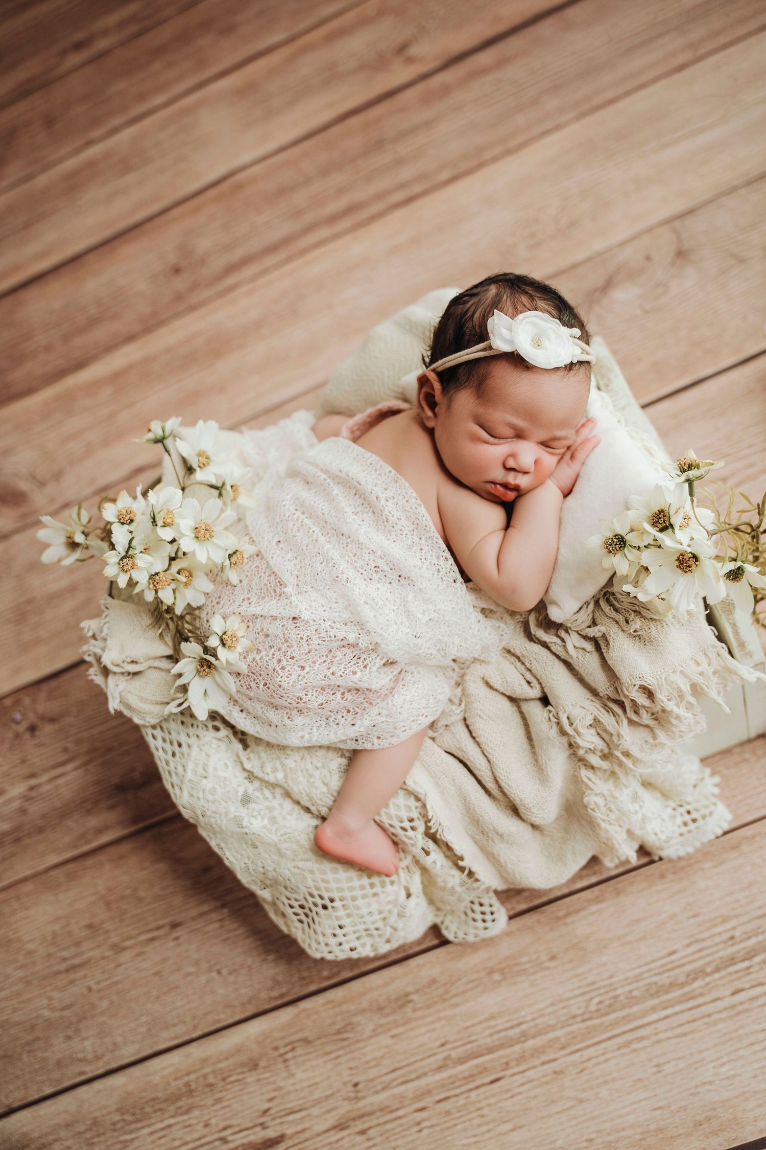 Sleeping newborn baby girl in white lace dress and floral headband resting on cream blanket with white flowers.