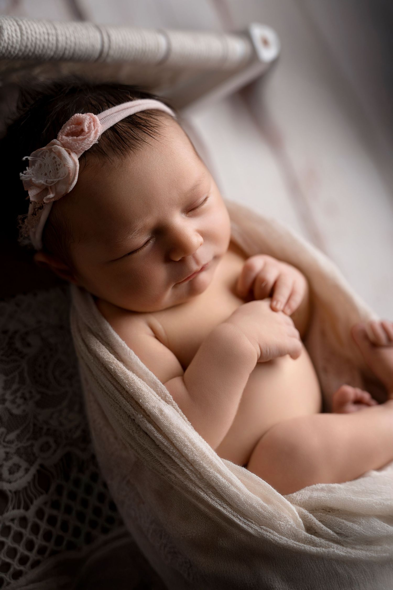 Serene newborn baby girl with pink floral headband sleeping in white lace bowl.