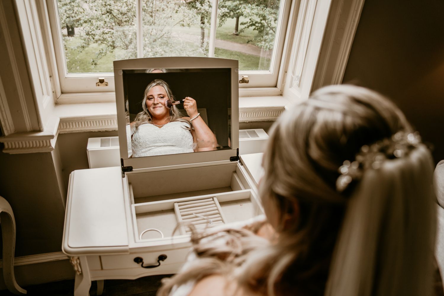Bride admires herself in vanity mirror wearing white gown and floral hair accessory.