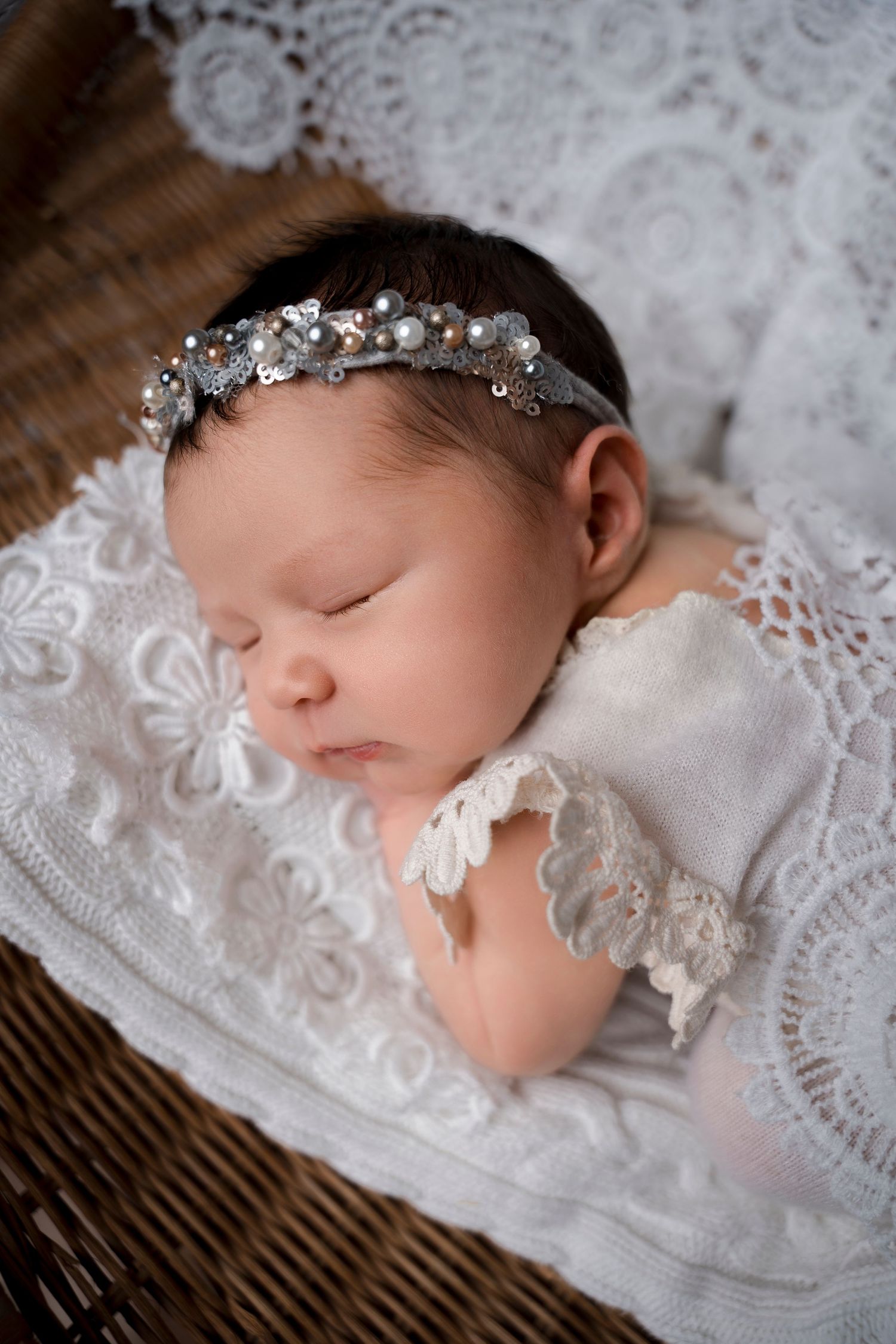 Sleeping newborn baby girl in silver beaded headband on white lace in wicker basket.