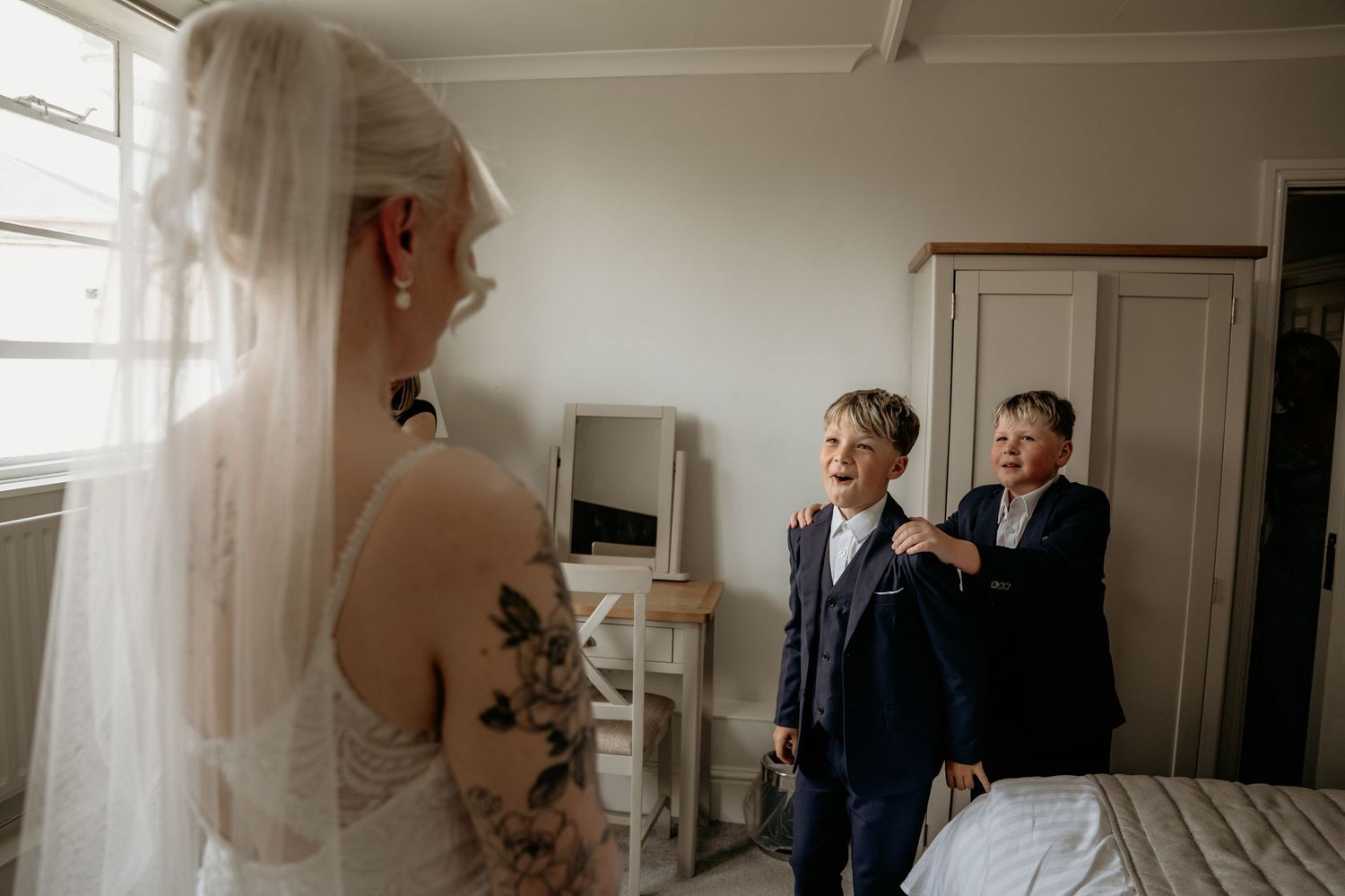 Bride in a lace gown surprises two young boys in suits in a bright bedroom before the ceremony.