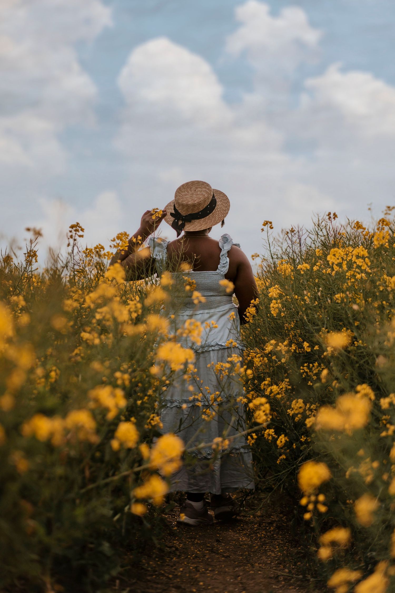 Woman in white dress and straw hat walking through yellow flower field.
