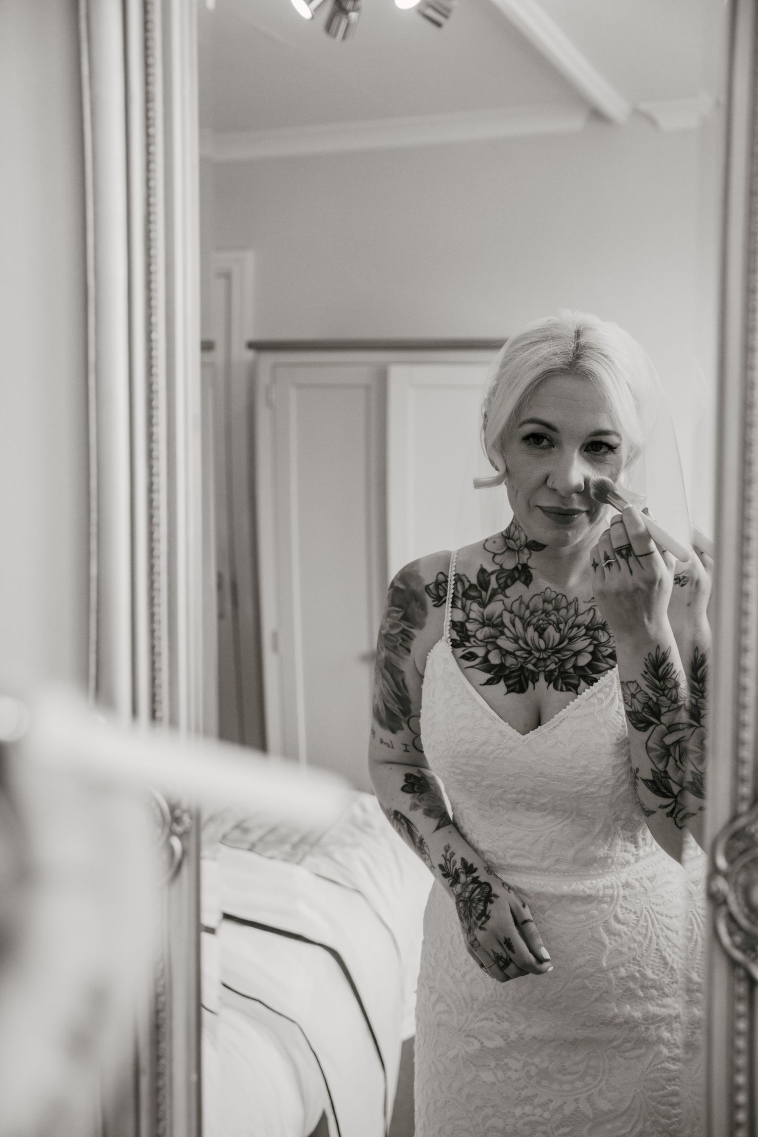 Heavily tattooed blonde bride in lace dress applies lipstick in elegant ornate mirror, black and white.