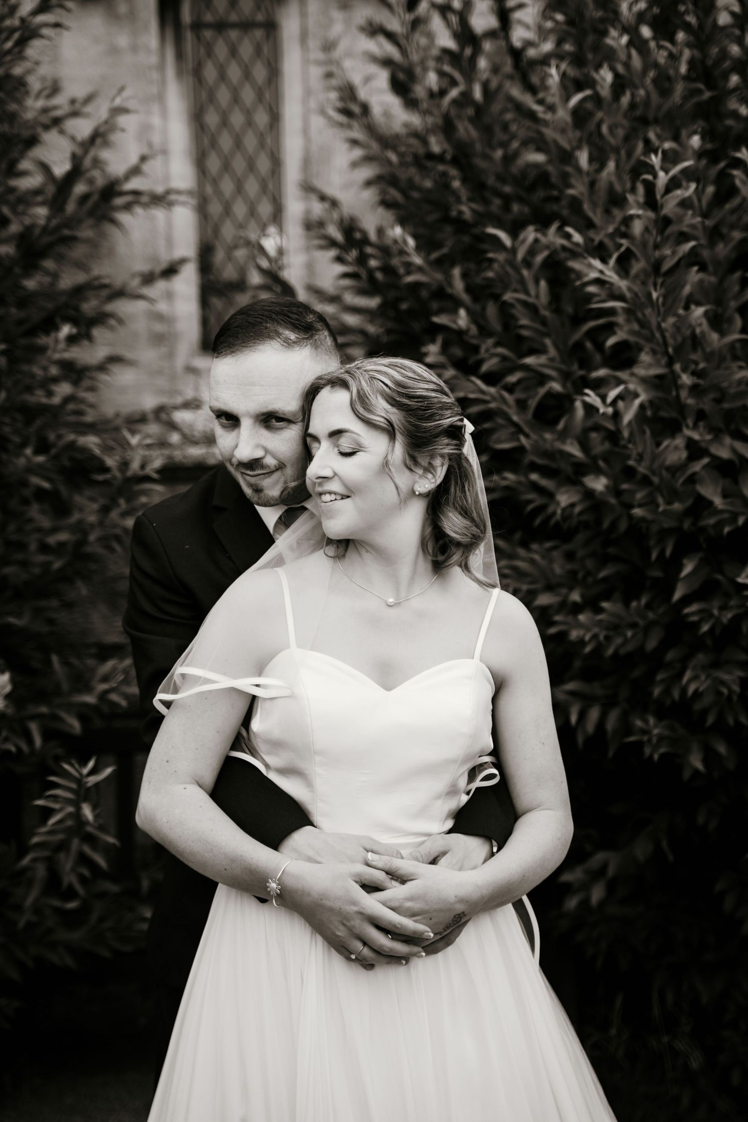 Groom tenderly embraces smiling bride in strapless gown among lush green foliage in black and white.