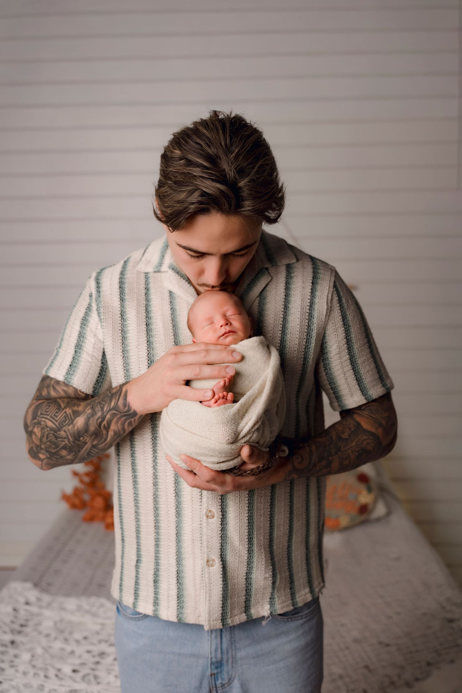 Tattooed young father in striped shirt gently kissing his swaddled newborn baby in a cozy studio setting.