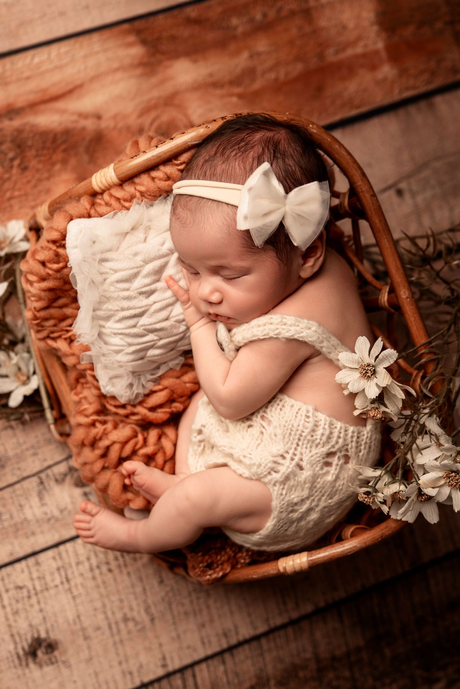 Newborn baby girl sleeping in a wicker basket with a white bow headband and cream outfit.