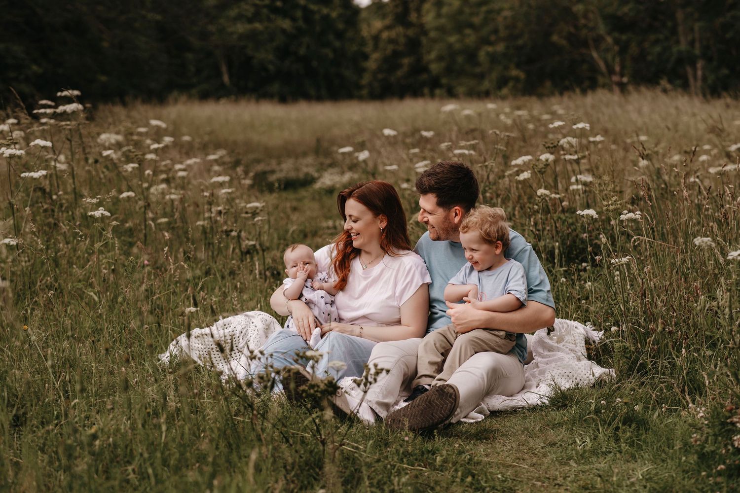 Family of four sitting on blanket in meadow, parents smiling with two young children surrounded by wildflowers.