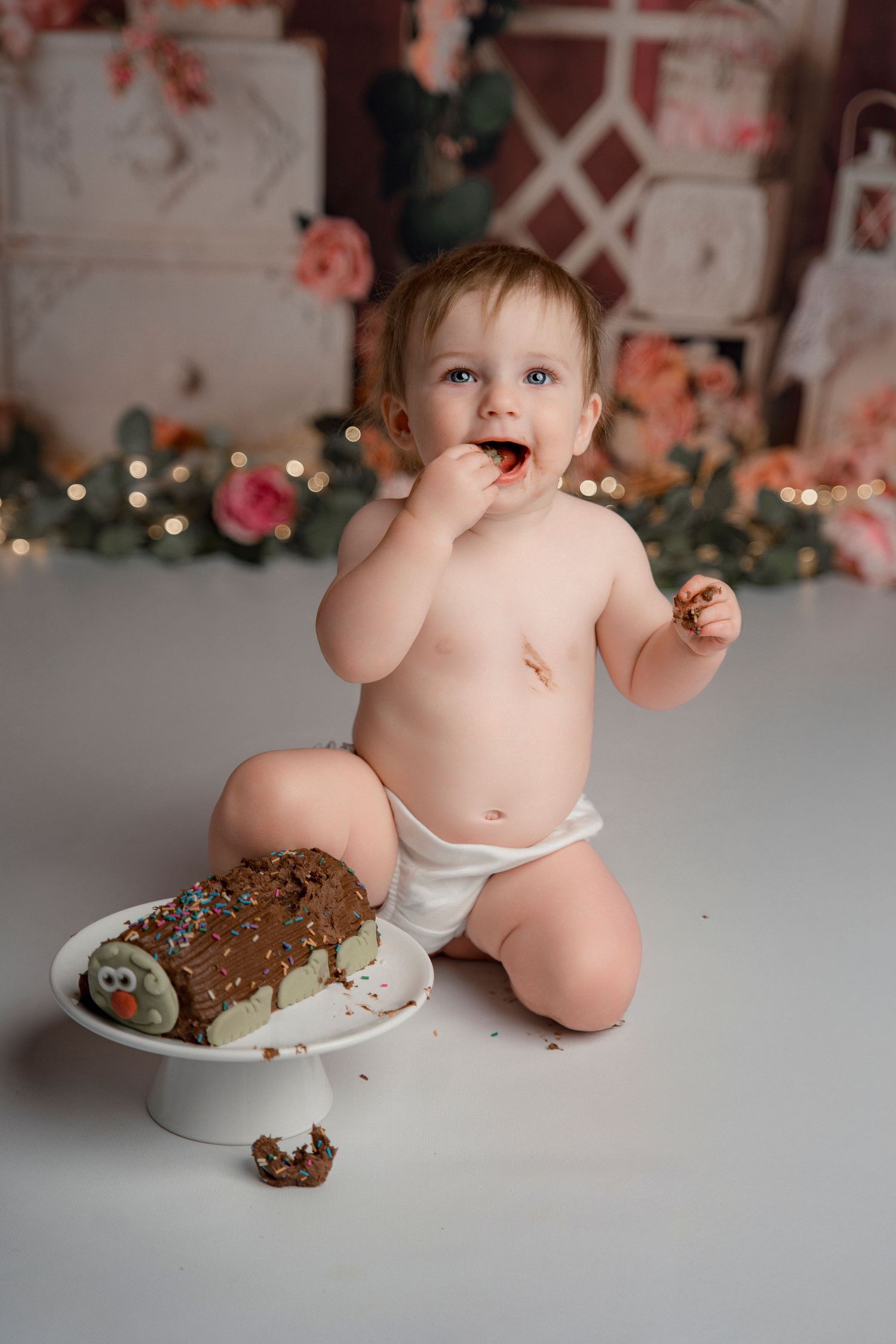 Baby in diaper eating chocolate cake during first birthday cake smash session with floral backdrop.