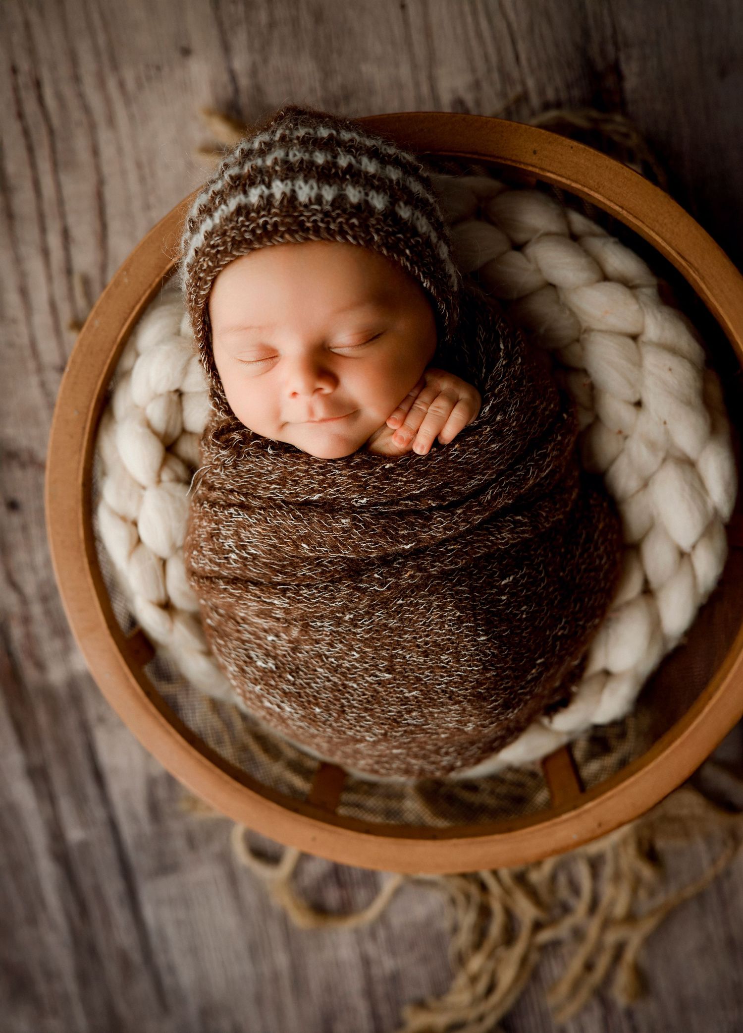 Newborn baby swaddled in brown knit wrap and striped bonnet sleeping in a wooden bowl.