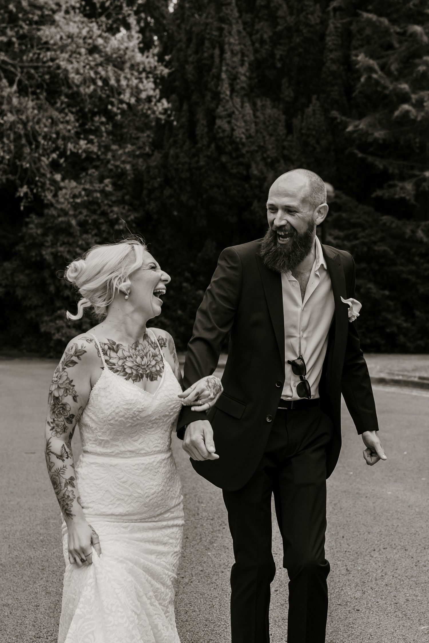 Laughing tattooed bride and bearded groom walking together in black and white photo.