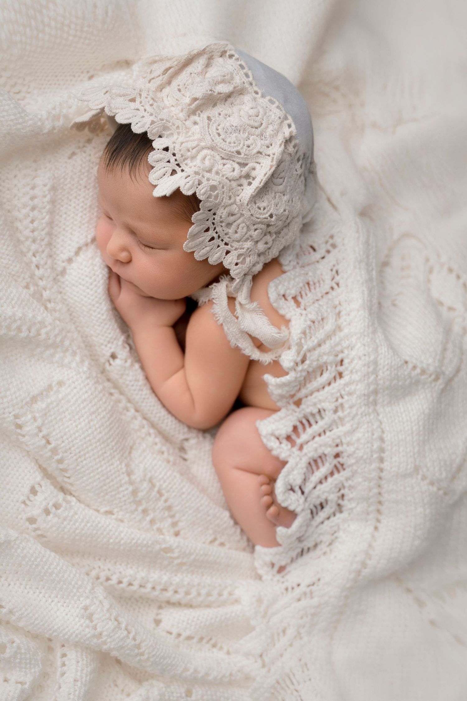 Peaceful newborn baby sleeping wrapped in white lace bonnet on knit blanket.