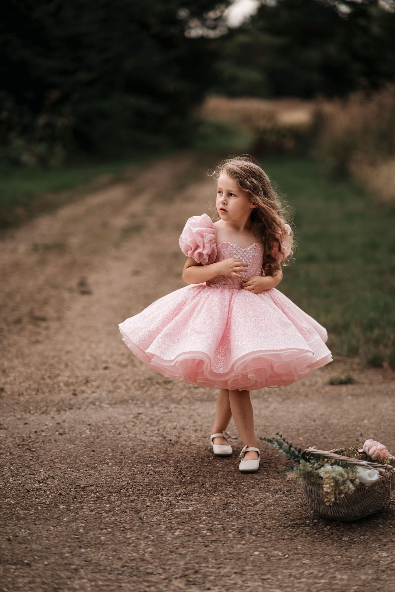 A little girl in a pink tulle dress stands on a dirt path holding flowers, with a basket of blooms beside her.