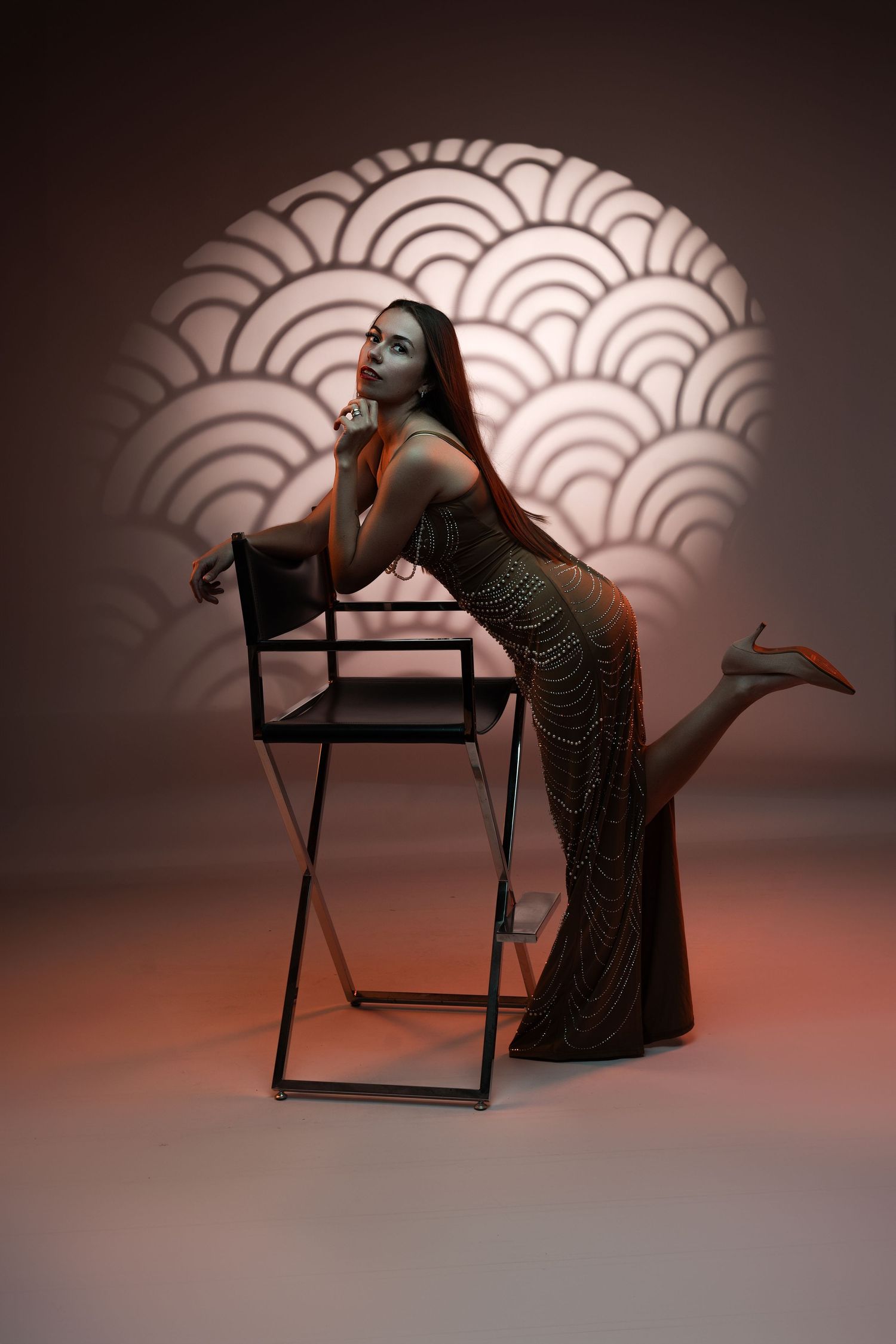 Elegant woman in a beaded gown posing beside a director's chair against a decorative art deco light pattern backdrop.