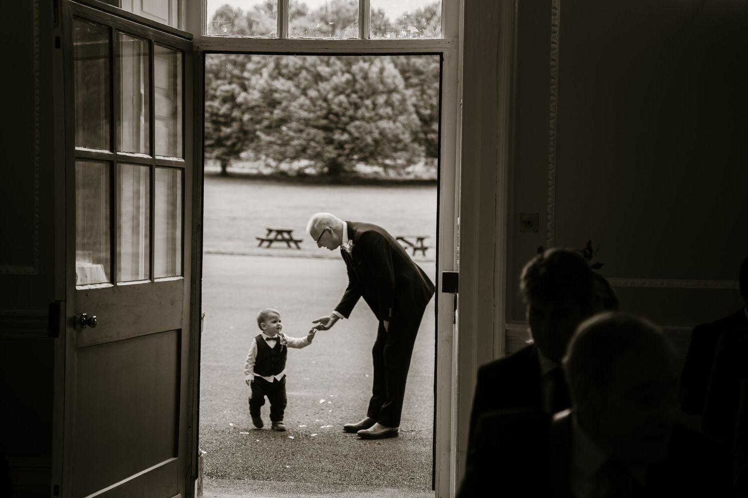 An elderly man in a suit bends down to greet a toddler in a doorway at a wedding.