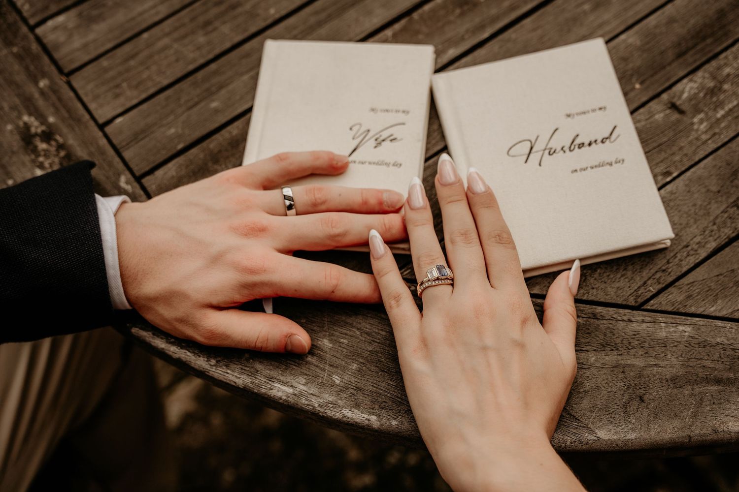 Two hands with wedding rings rest on a wooden table beside marriage booklets, symbolizing a romantic union.