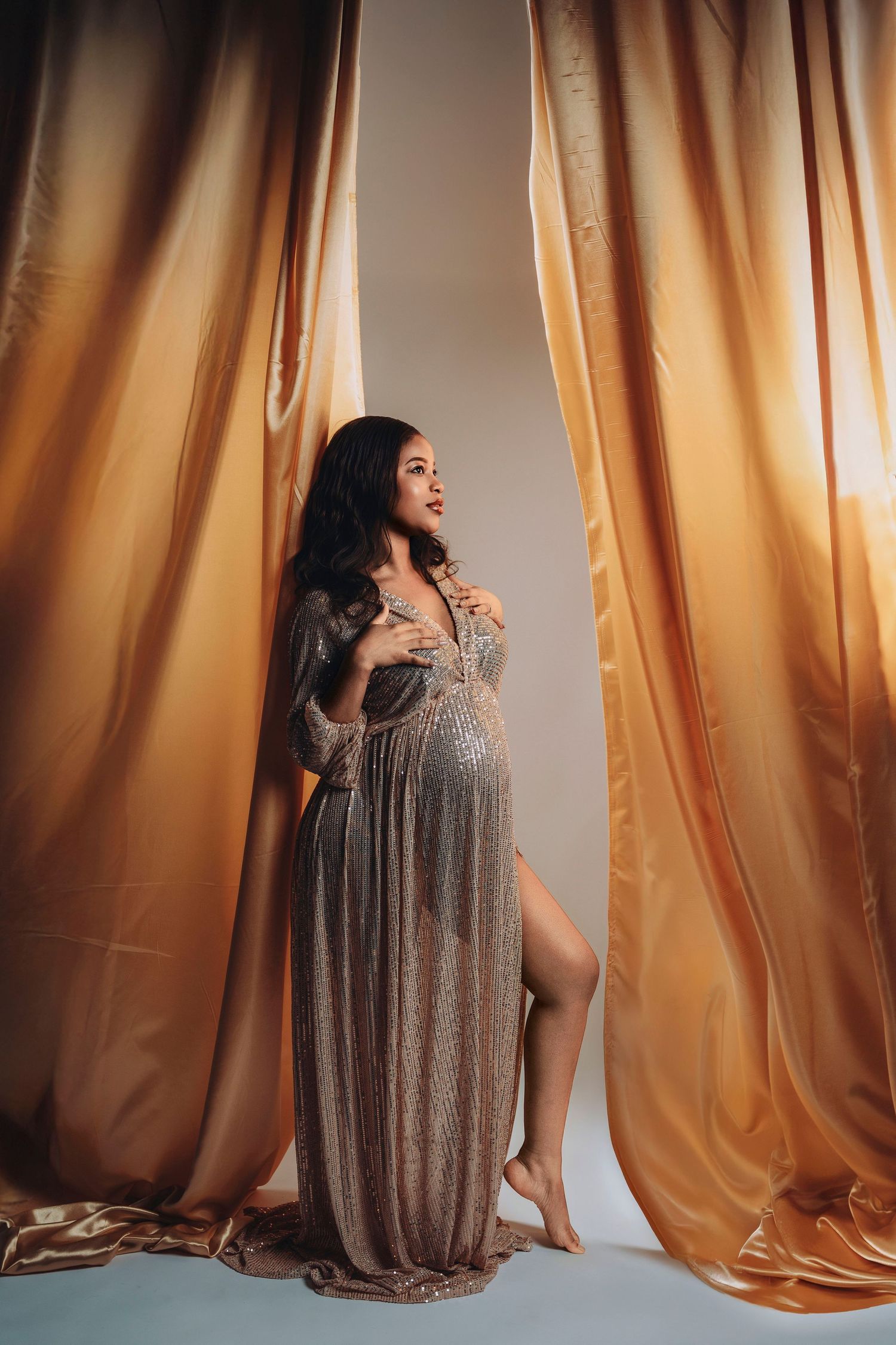 Pregnant woman posing elegantly in a shimmering gold maternity gown between golden satin curtains in a studio.