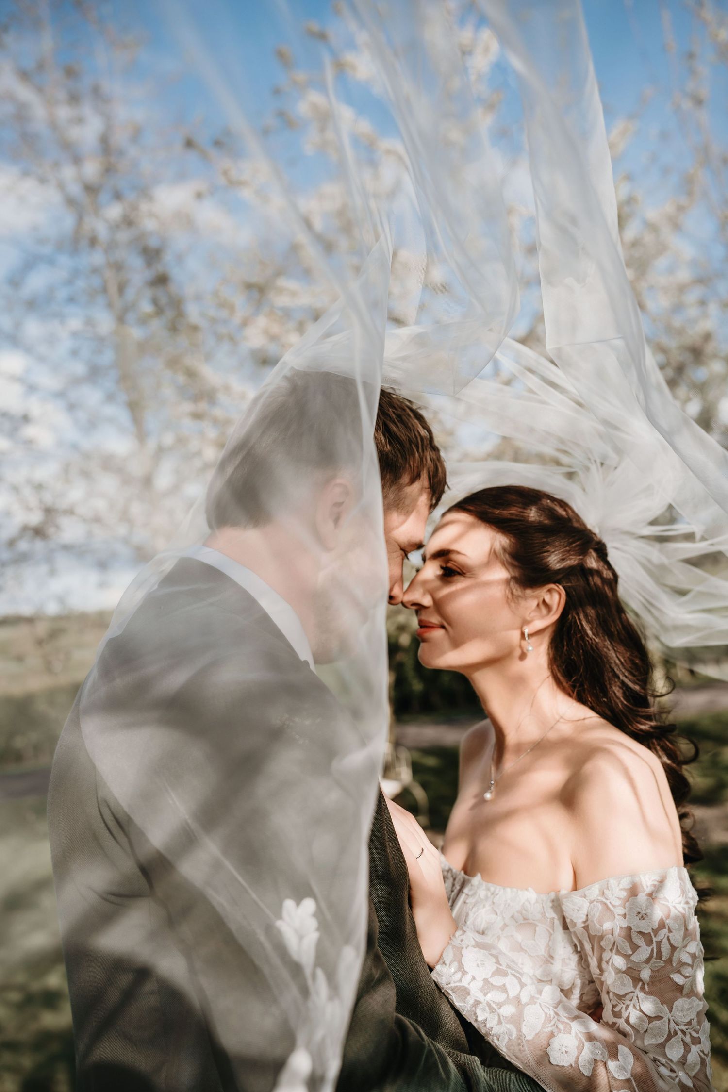 Bride and groom share a romantic moment under a flowing veil with blooming trees in the background.