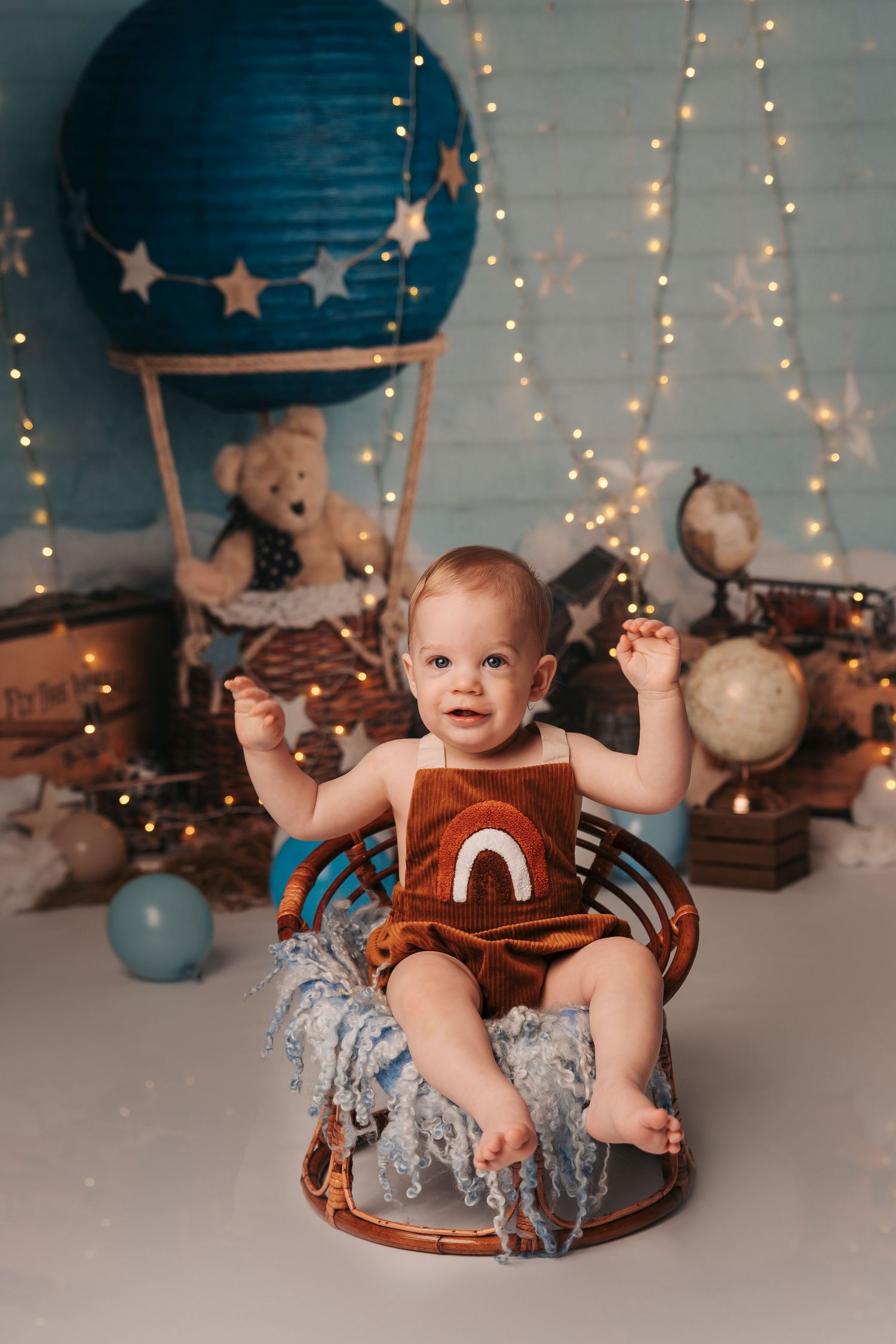 Baby sitting in wicker chair with rainbow book, surrounded by hot air balloon decor and fairy lights.