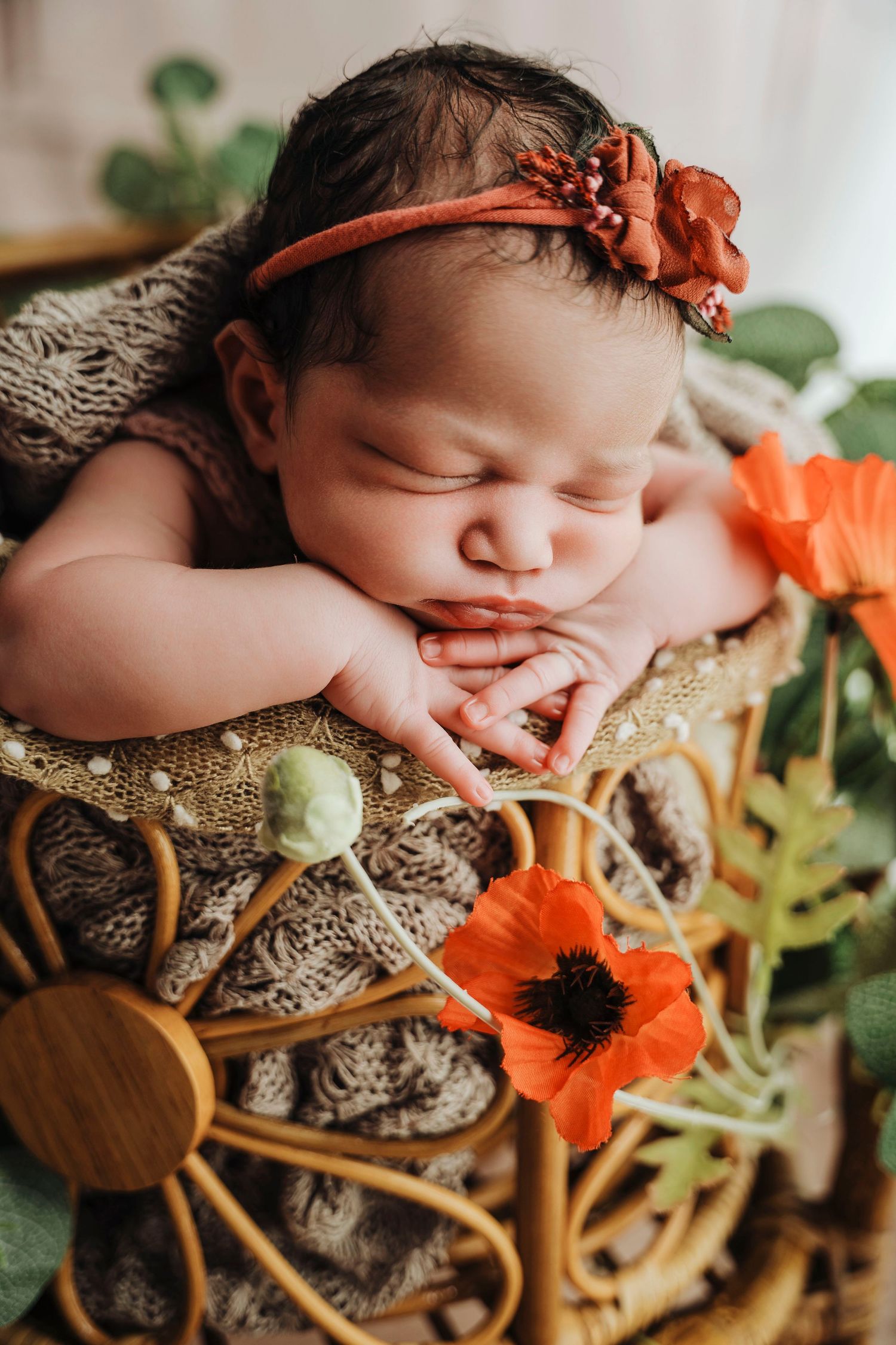 Sleeping newborn baby with orange floral headband resting in a wicker basket surrounded by red poppy flowers.