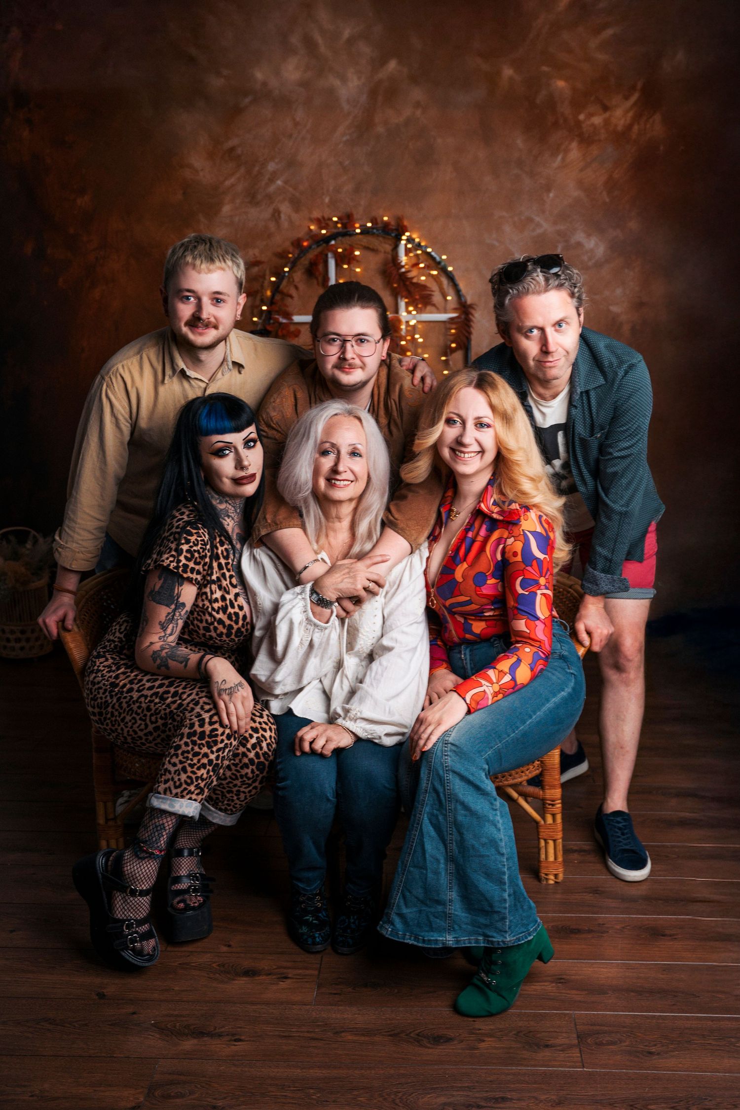 Group of six adults posing together for a studio portrait with warm brown backdrop and decorative wreath with lights.