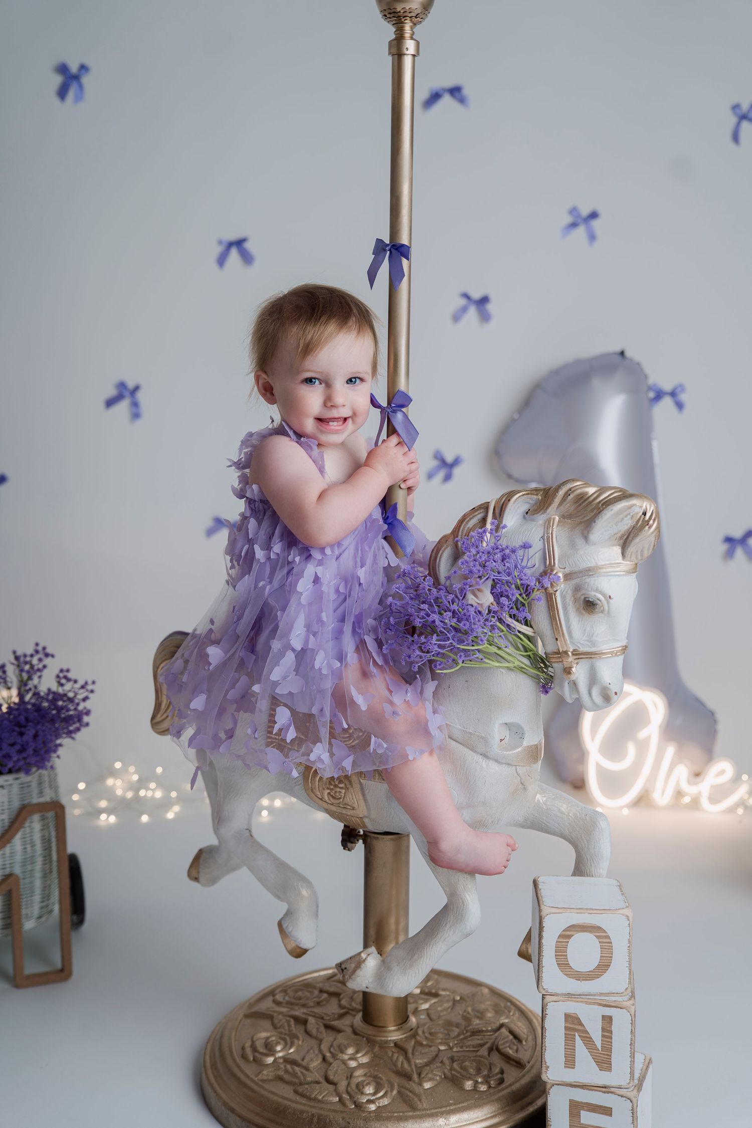 Smiling toddler girl in purple tulle dress sitting on white carousel horse with lavender flowers and ONE birthday decor.