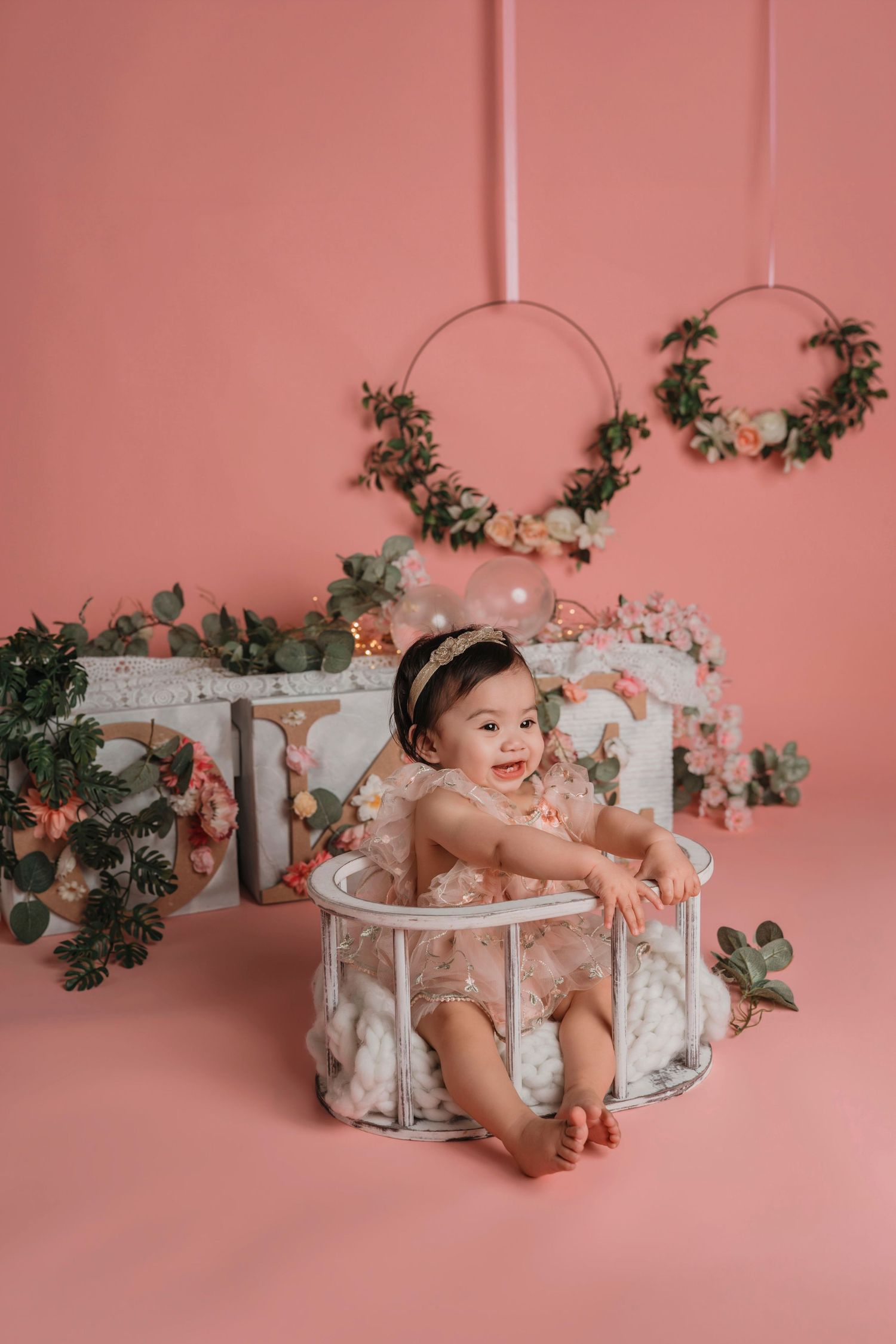 Smiling baby girl in lace outfit sits in decorative basket amid pink floral backdrop with wreath decorations.