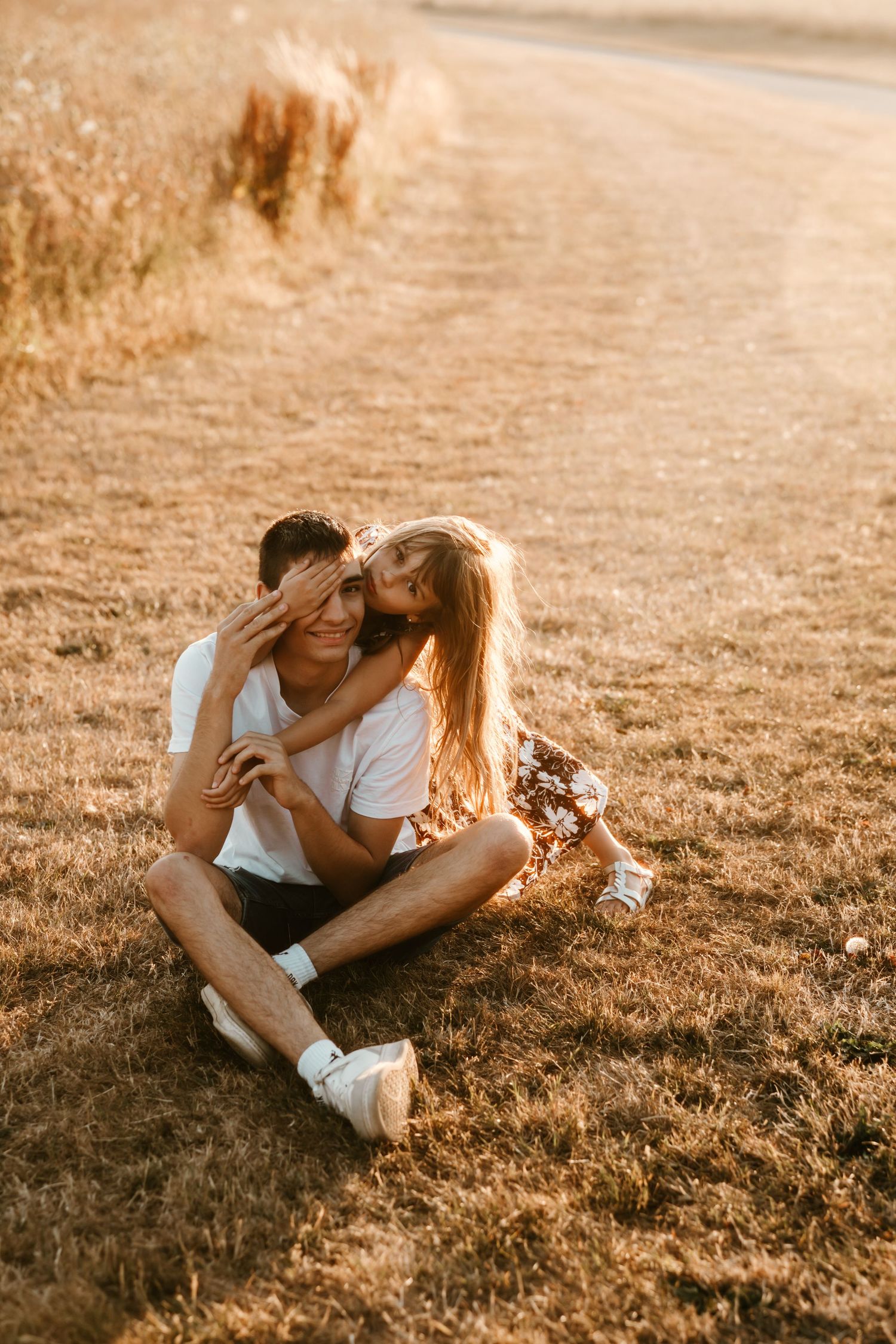 Couple sitting on golden grass at sunset, woman kissing man on cheek while covering his eyes romantically.