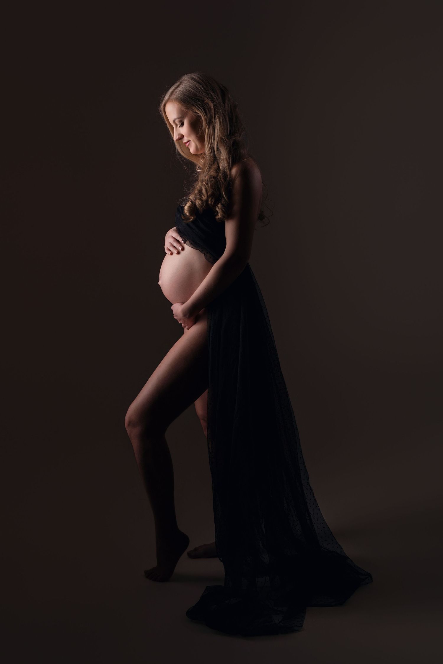 Pregnant woman in black sheer fabric posing on dark background.