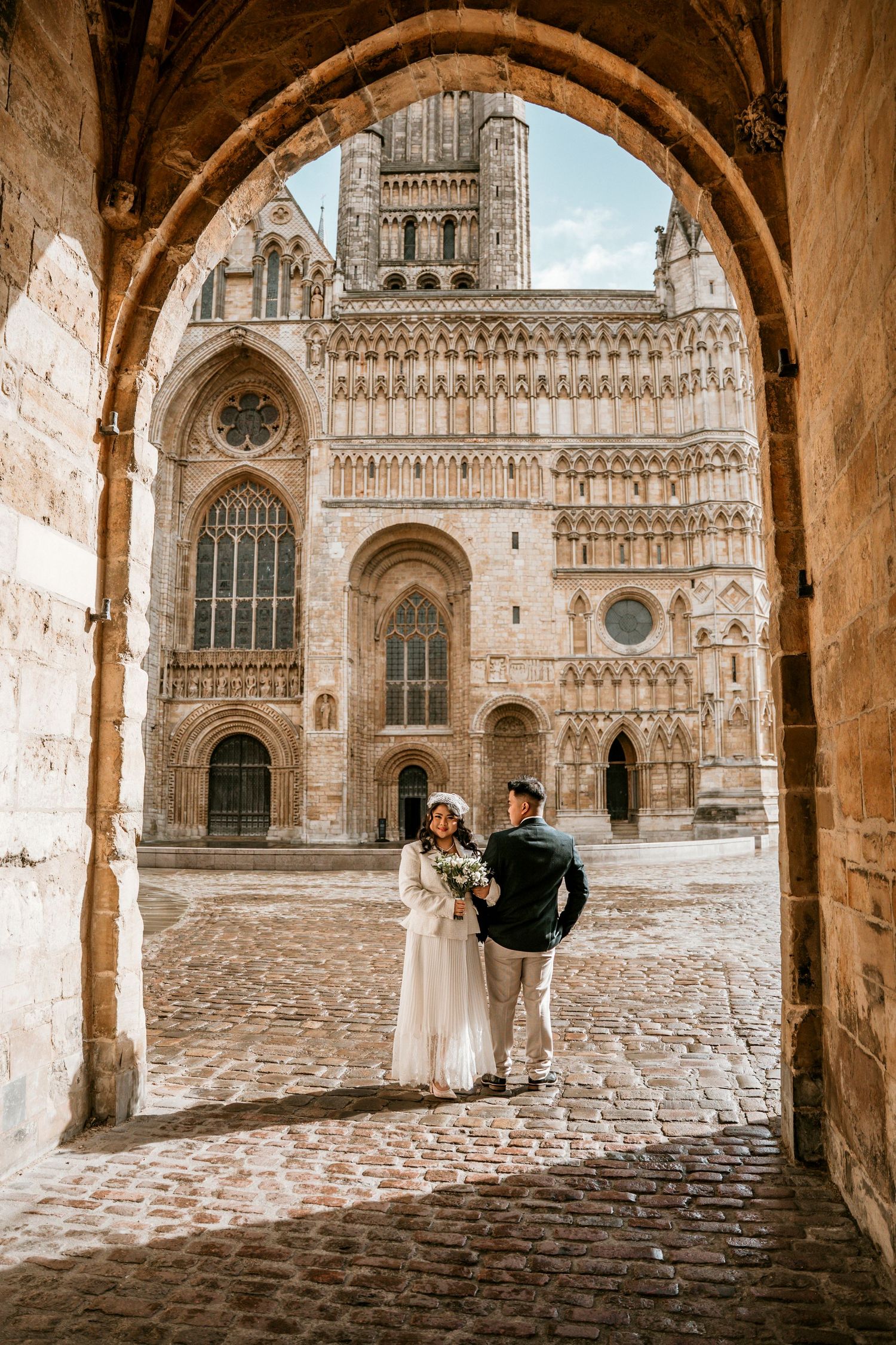 Bride and groom stand under stone arch before ornate Gothic cathedral on cobblestone plaza.