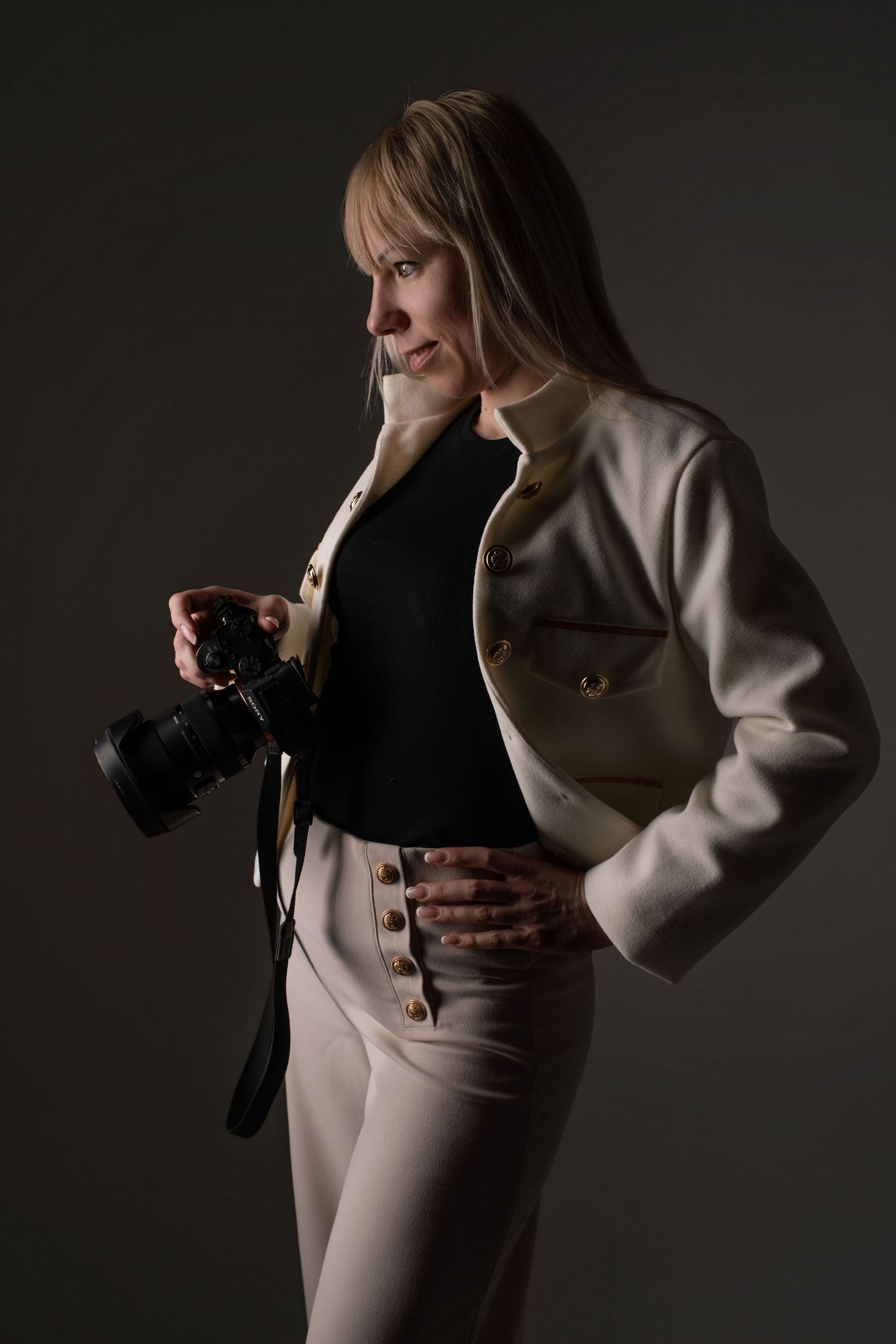 Professional female photographer in a white blazer holds a camera while posing in a dark studio.