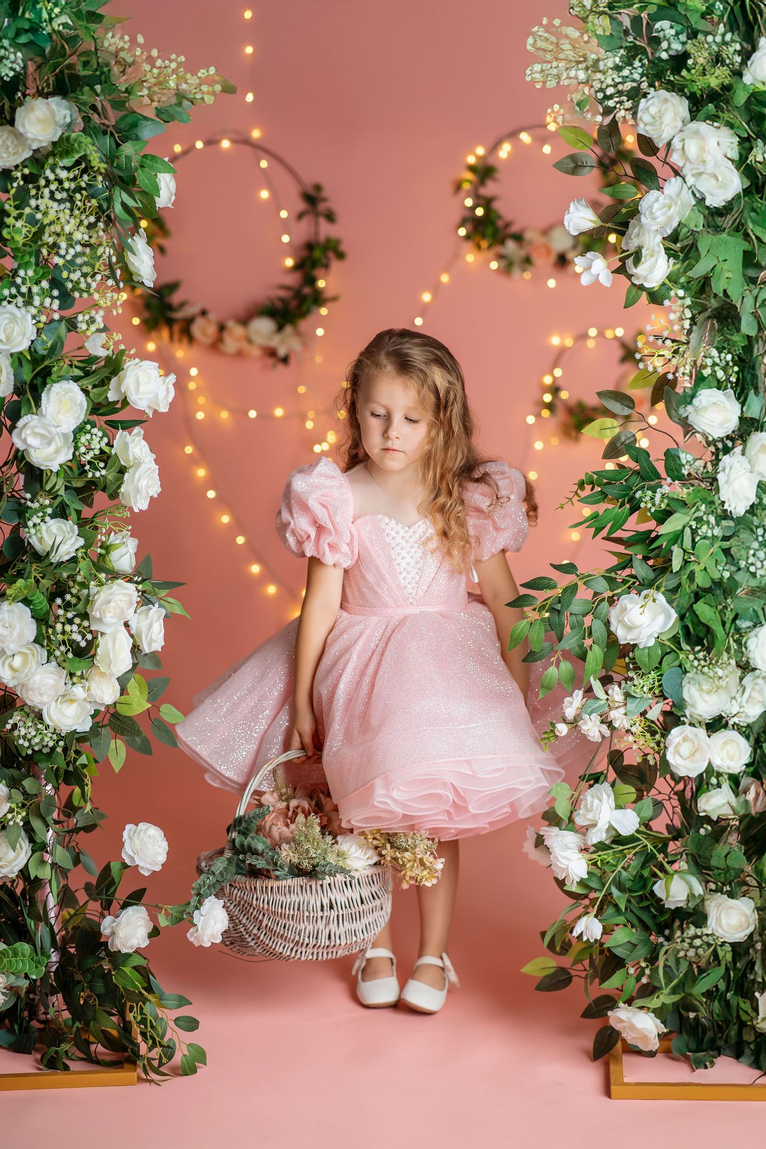 Young girl in pink dress holding basket among white floral arches with fairy lights on pink background.