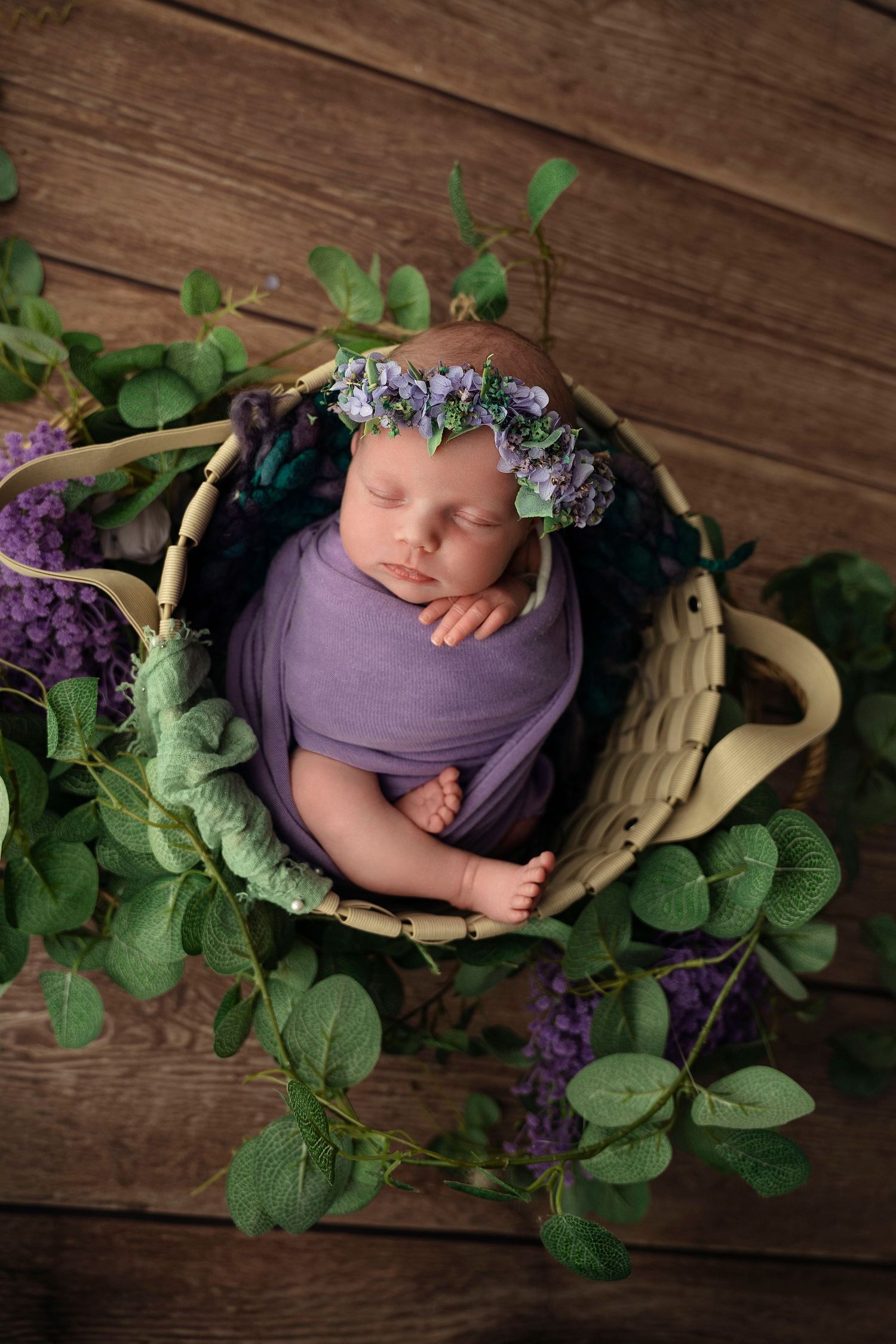 Sleeping newborn wrapped in purple swaddle with floral crown in wicker basket.