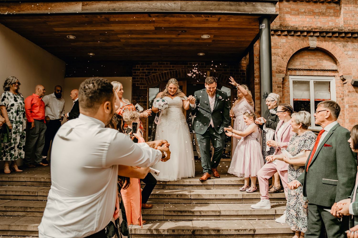 Joyful newlyweds exit venue through confetti shower surrounded by celebrating wedding guests on steps.